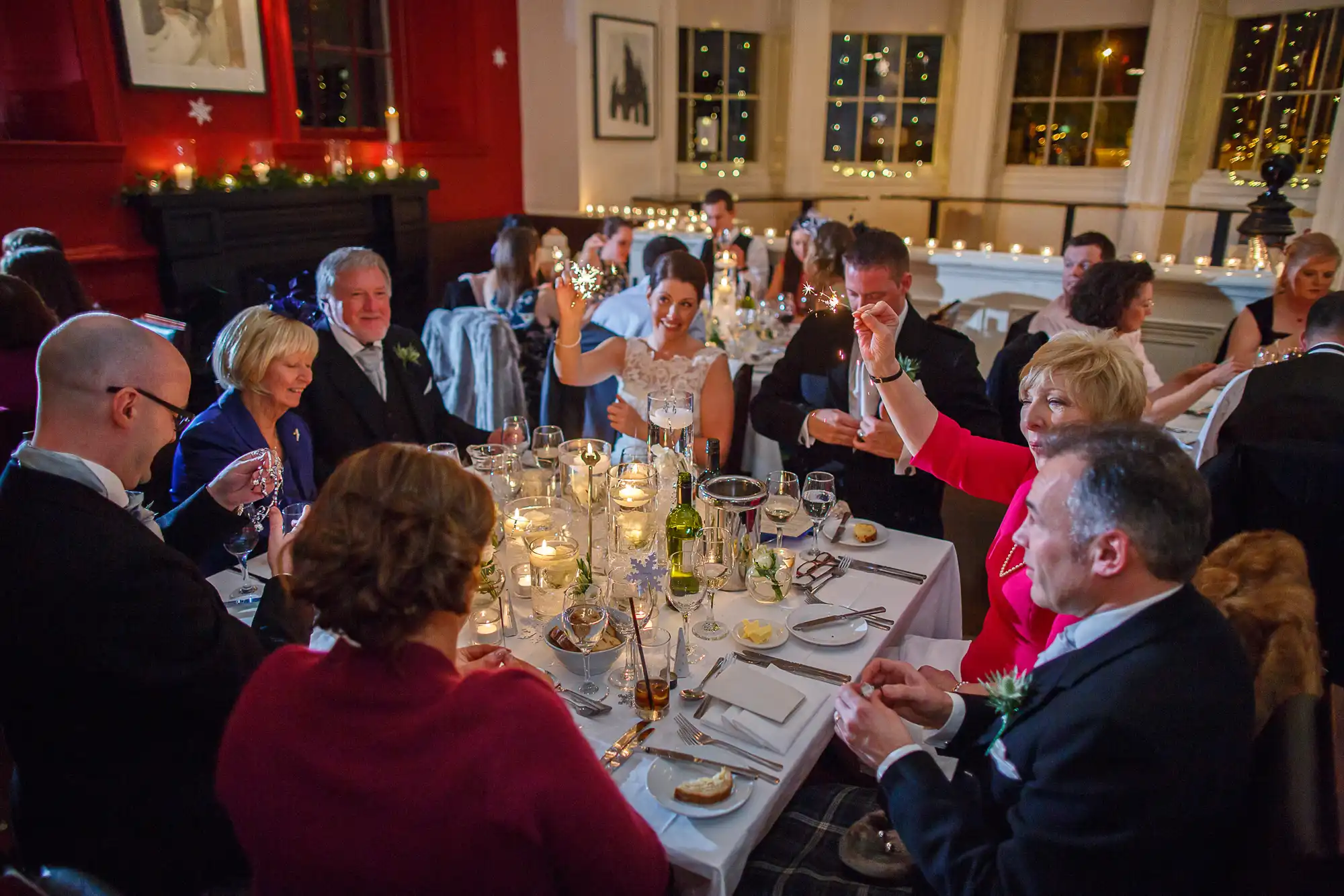 People enjoying a dinner party in a festive, candlelit room, raising glasses in a toast, with holiday decorations visible.