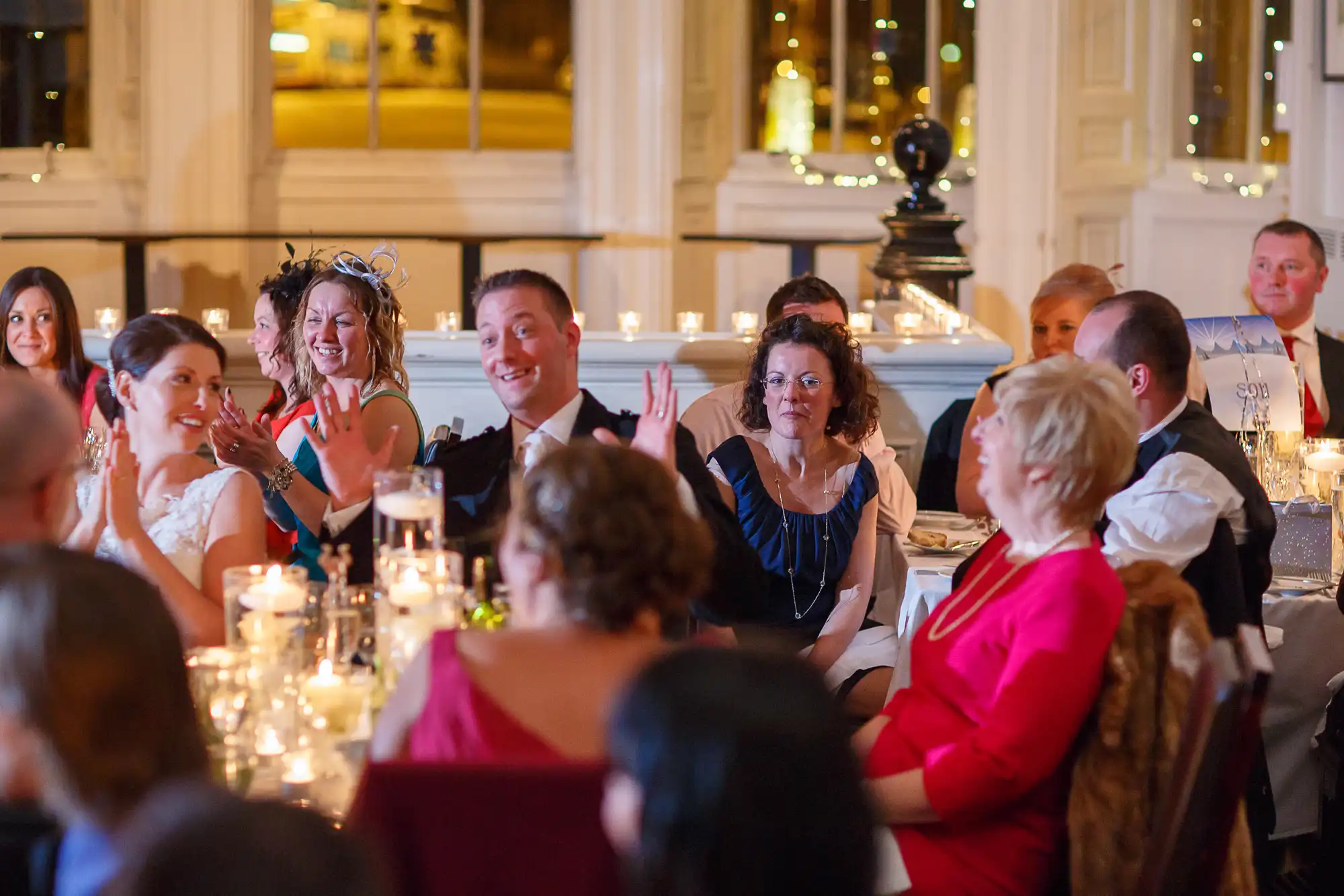 Wedding guests seated at ornately decorated tables, listening and reacting to speeches in a warmly lit banquet hall.