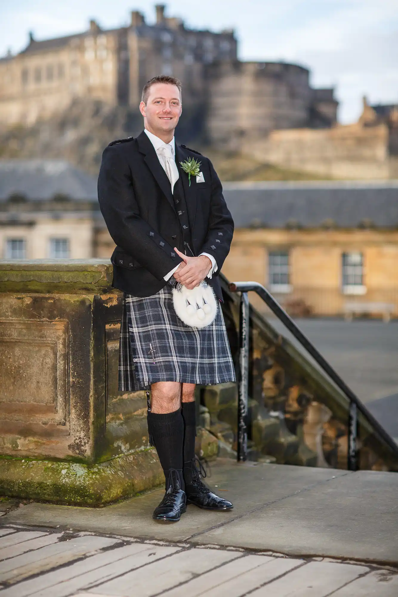 Man in a traditional scottish kilt and jacket, smiling, standing on a cobblestone path with edinburgh castle in the background.
