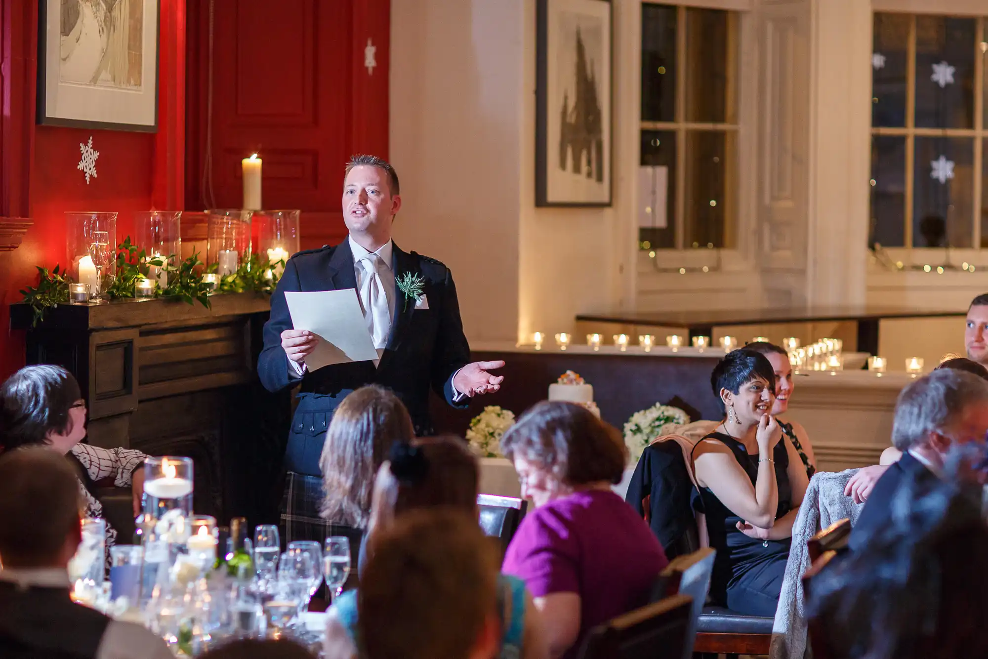 Man in suit giving a speech at a wedding reception, guests seated around tables listening, room decorated with candles and lights.
