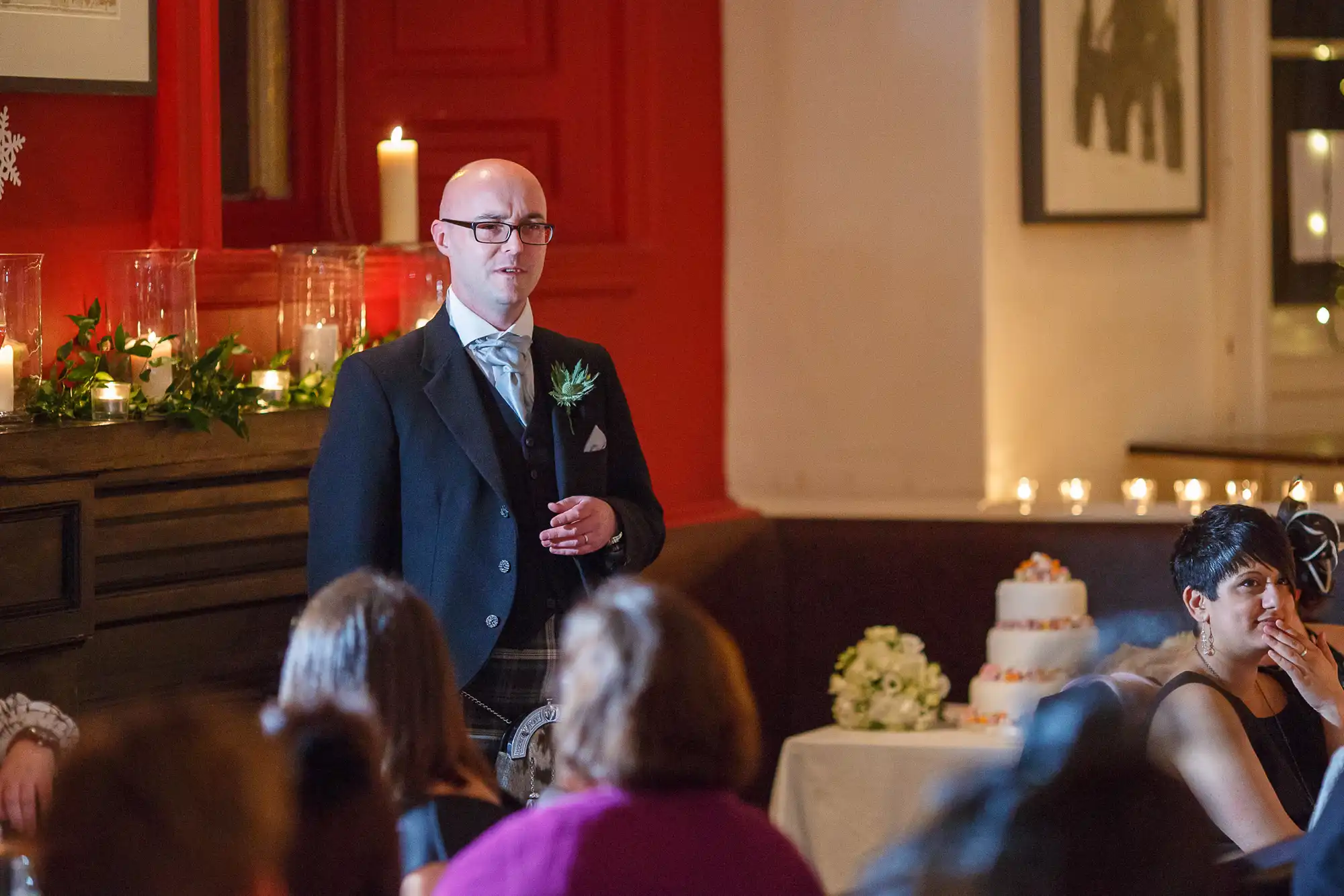 A bald man in a suit giving a speech at a wedding reception, holding a microphone, with a wedding cake and guests in the background.