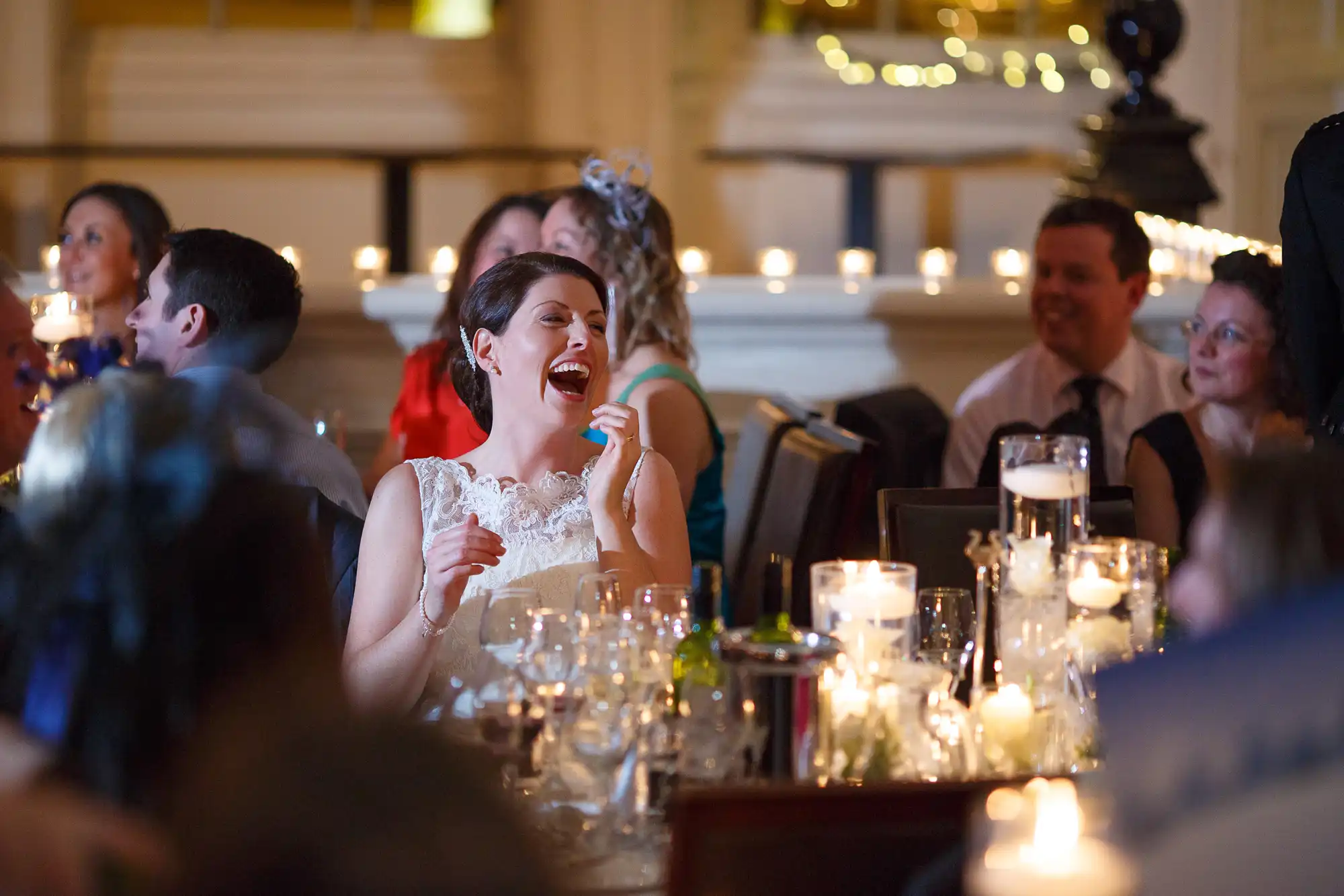 A bride laughing joyfully at a candlelit reception table surrounded by guests.