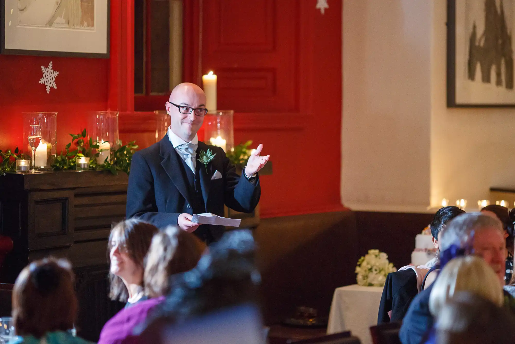 A man in a suit giving a speech at a candlelit event, guests seated around, in a room with red walls and festive decorations.