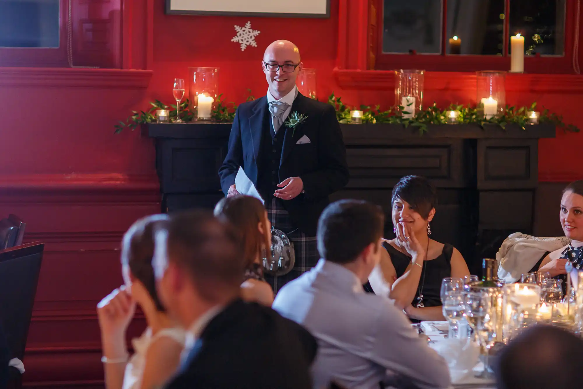 Man giving a speech at a wedding reception in a room with red walls, lit candles, and guests listening at dinner tables.