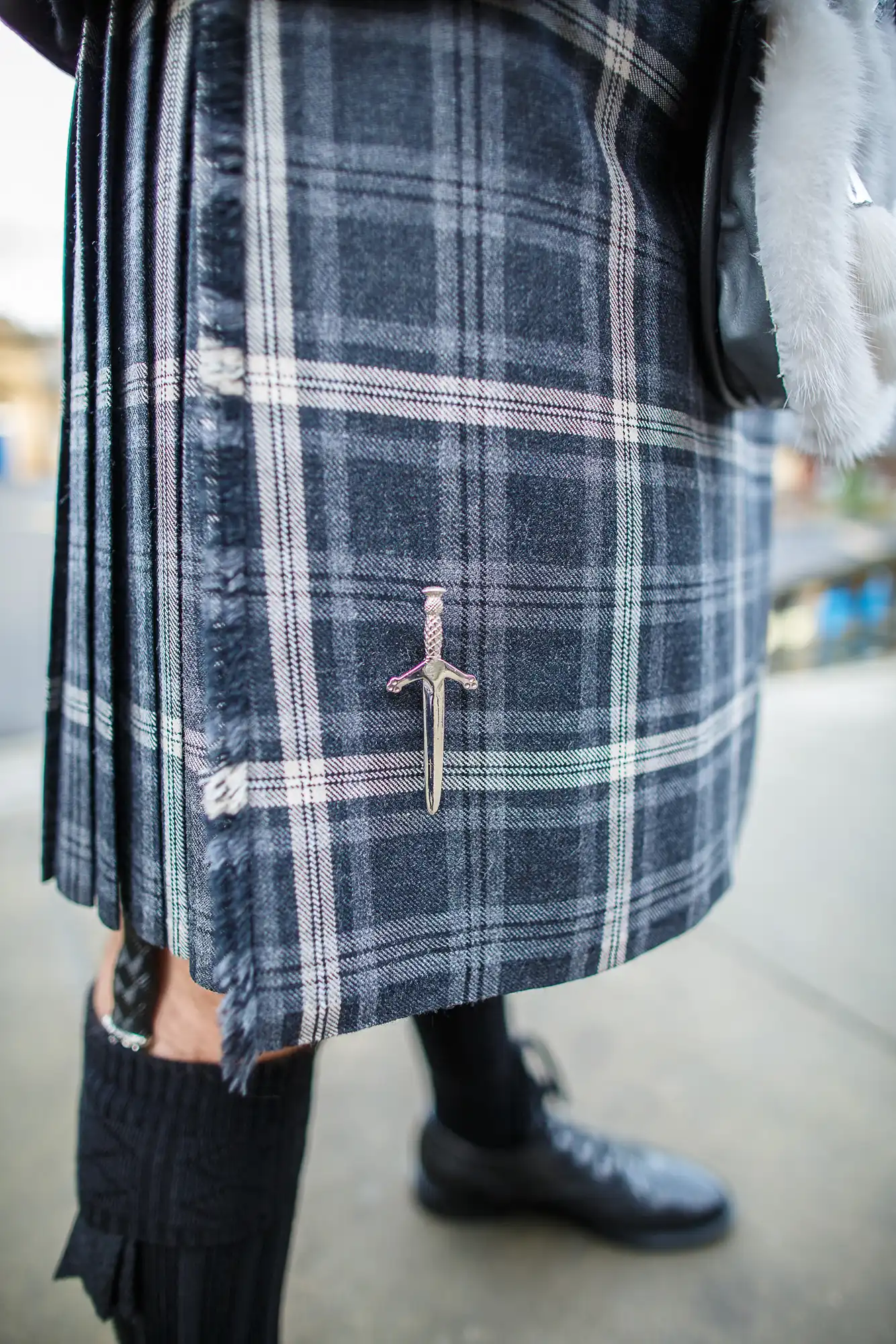 Close-up of a plaid skirt with a decorative sword-shaped kilt pin on it, complemented by black tights and shoes.