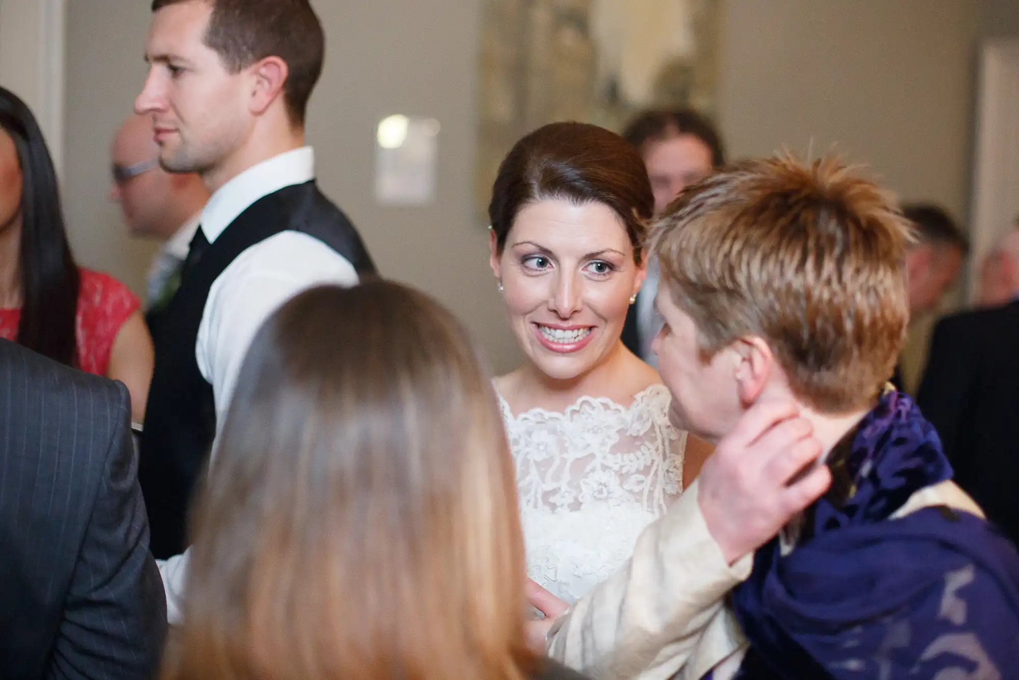 A smiling bride in a lace dress converses with guests at a wedding reception, with other attendees visible around her.