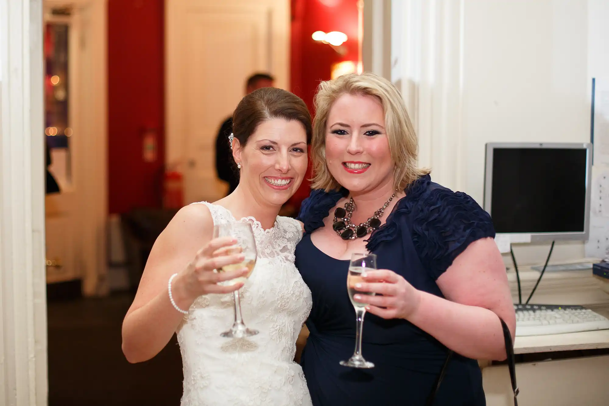 Two women smiling and toasting with champagne glasses at an indoor event, one dressed in a white wedding gown and the other in a dark blue dress.