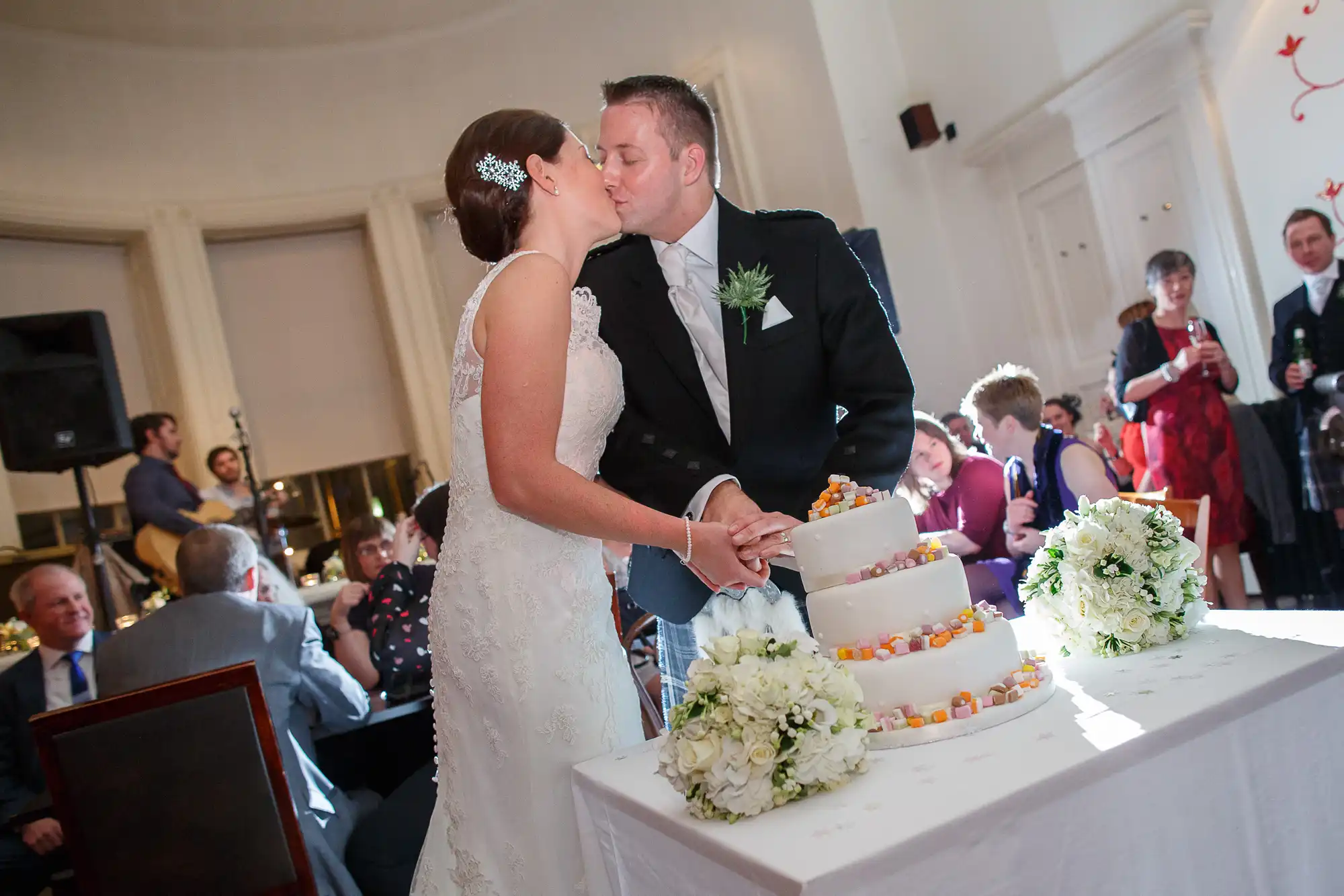 A bride and groom kiss while cutting a white tiered wedding cake, surrounded by guests in a brightly lit room.