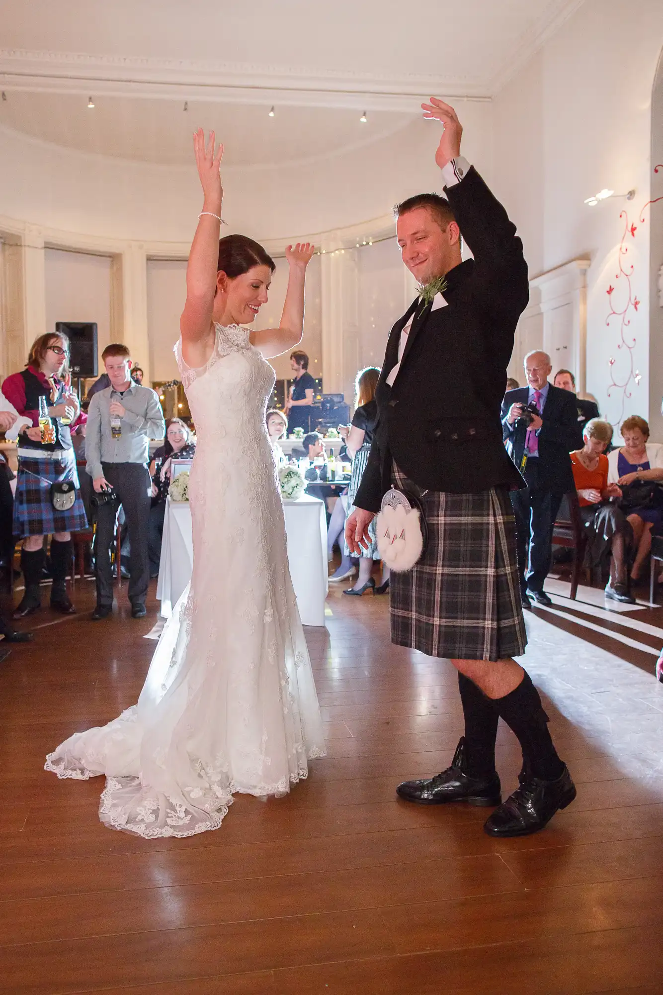 A bride in a white dress and a groom in a kilt joyfully dance with raised arms at their wedding reception, surrounded by guests.