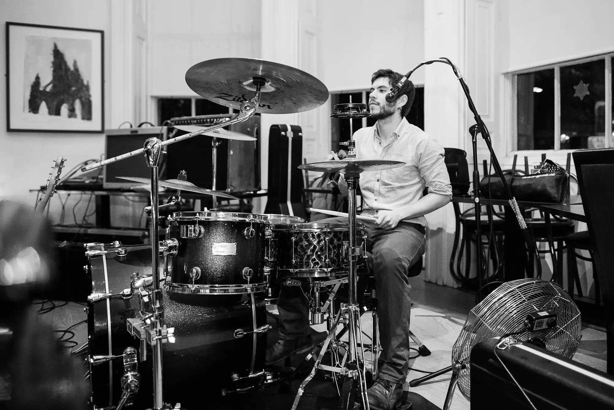 A male drummer playing a drum set in a room with large windows and artworks on the wall, captured in black and white.