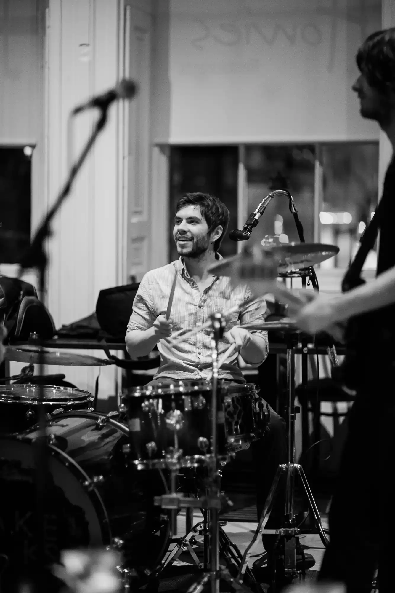 A smiling drummer sitting at his drum set, engaging with bandmates in a monochrome indoor setting.