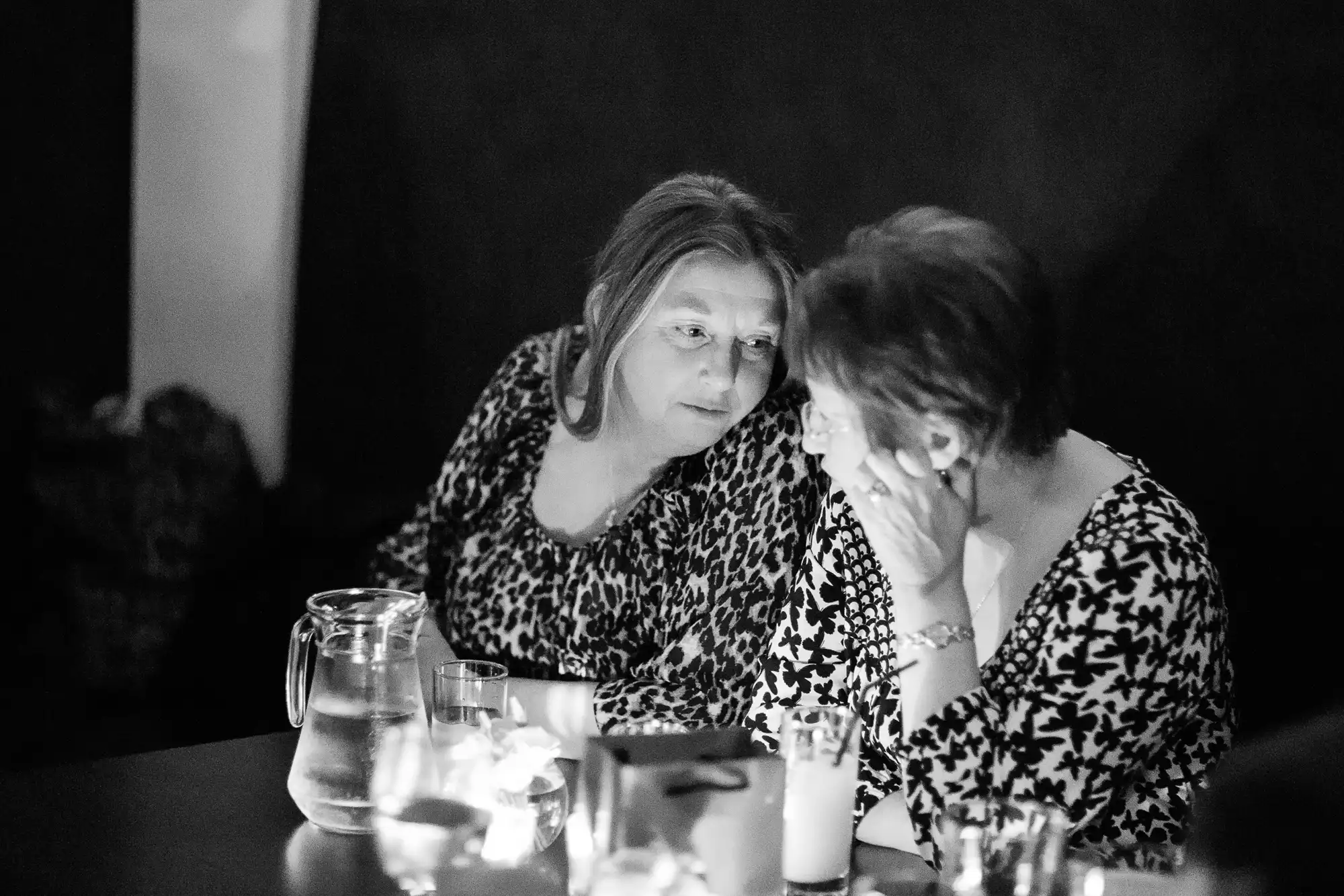 Two women engaged in a serious conversation at a dimly lit table with candles and a water pitcher.