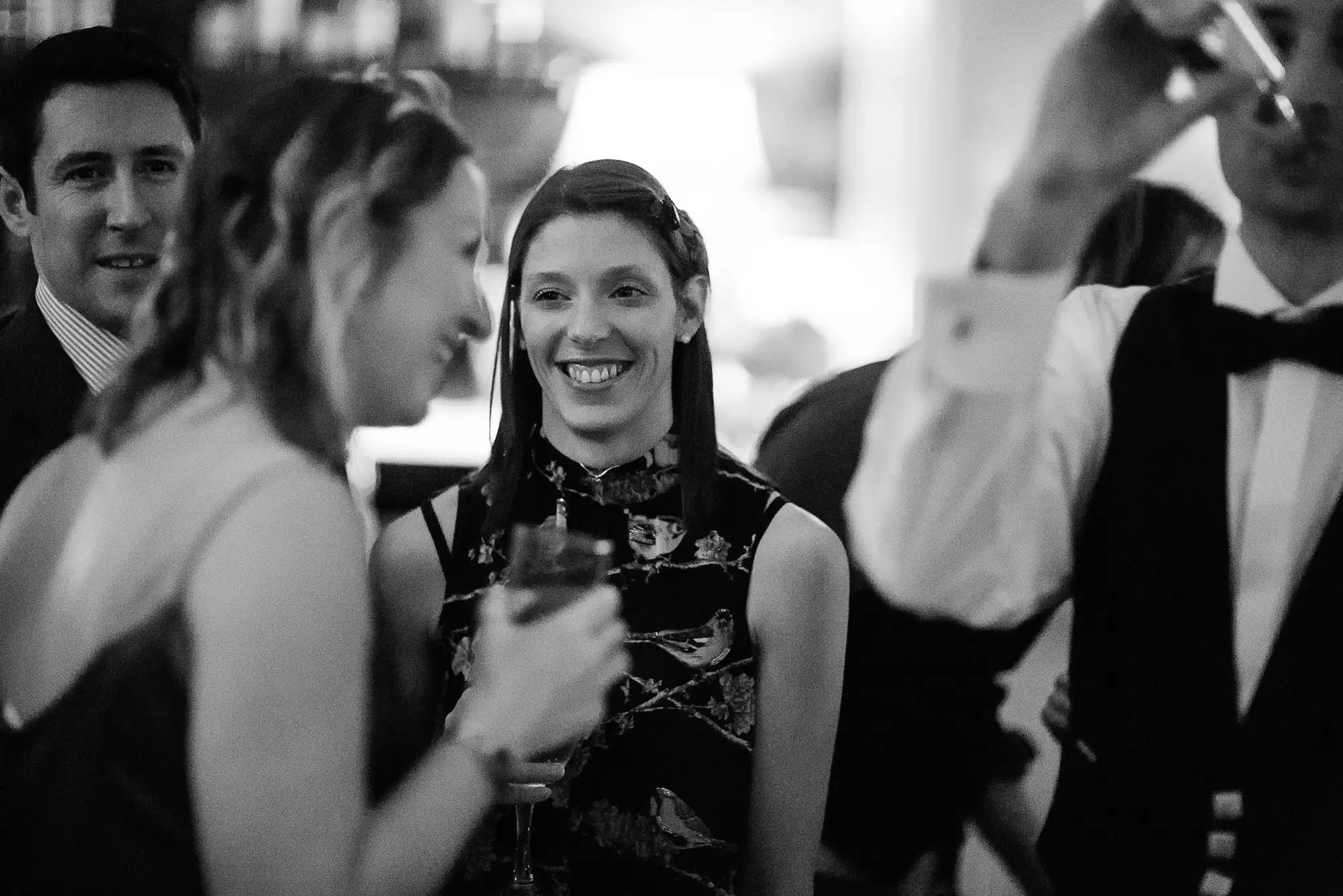 A woman with a ponytail smiles while holding a drink at a lively event, surrounded by other guests chatting in a dimly lit room.