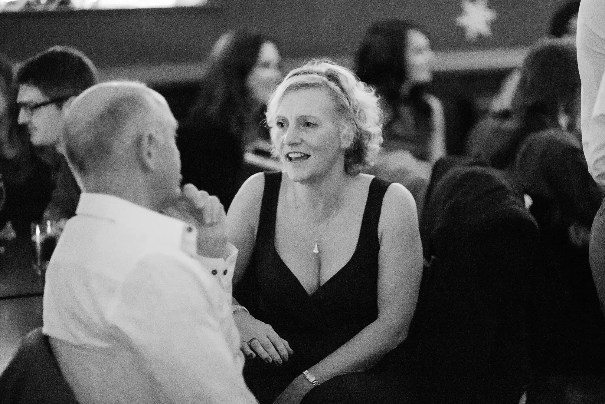A woman in a black dress converses with a man at a social gathering, both seated, in a black and white photo.