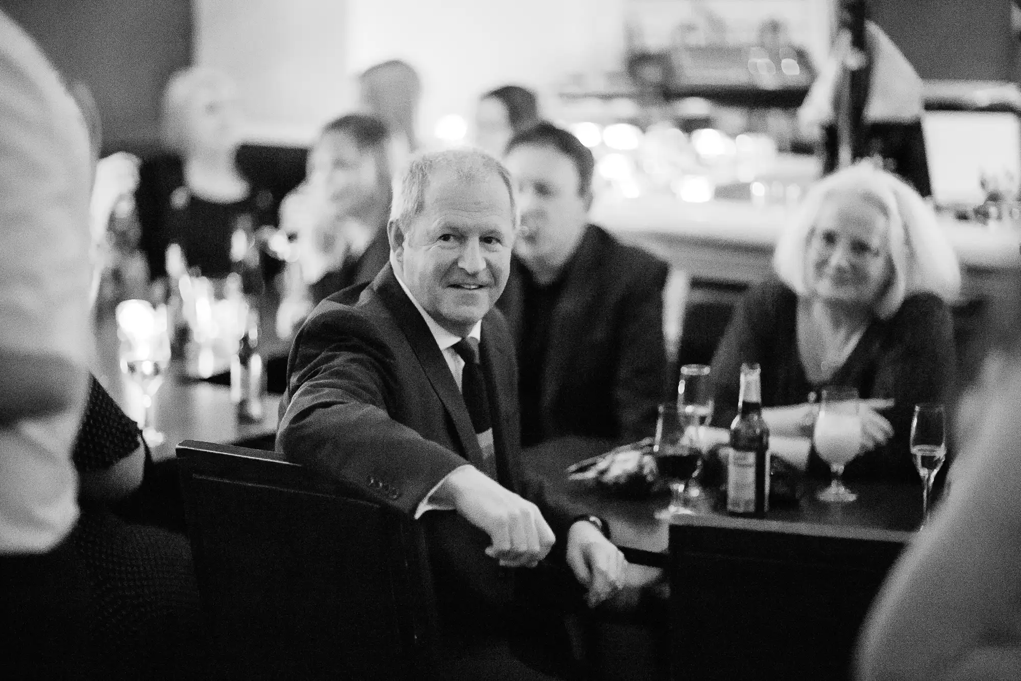 A man in a suit sitting at a bar, smiling and looking at the camera, with other people in the background.