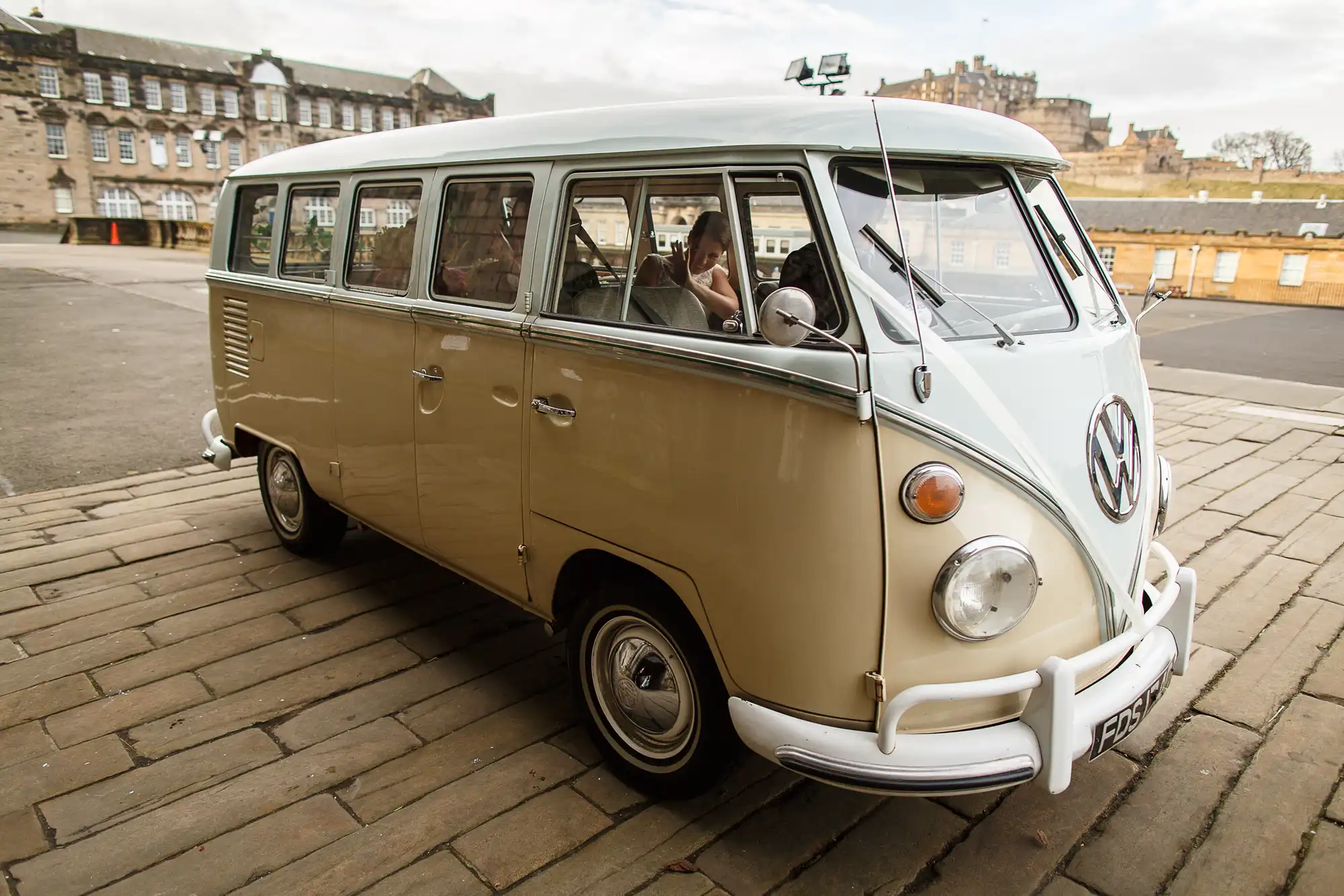 A vintage beige volkswagen van parked on a cobblestone street with a woman sitting in the driver's seat and a castle visible in the background.