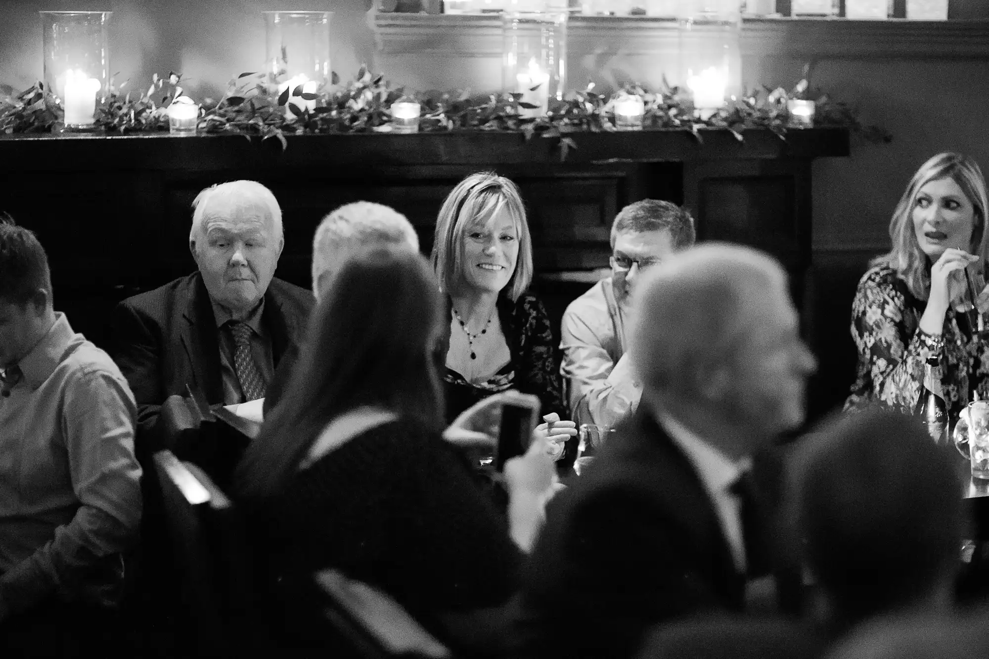 Black and white image of people at a crowded event, seated around tables, engaged in conversation with a background of dimly lit candles.