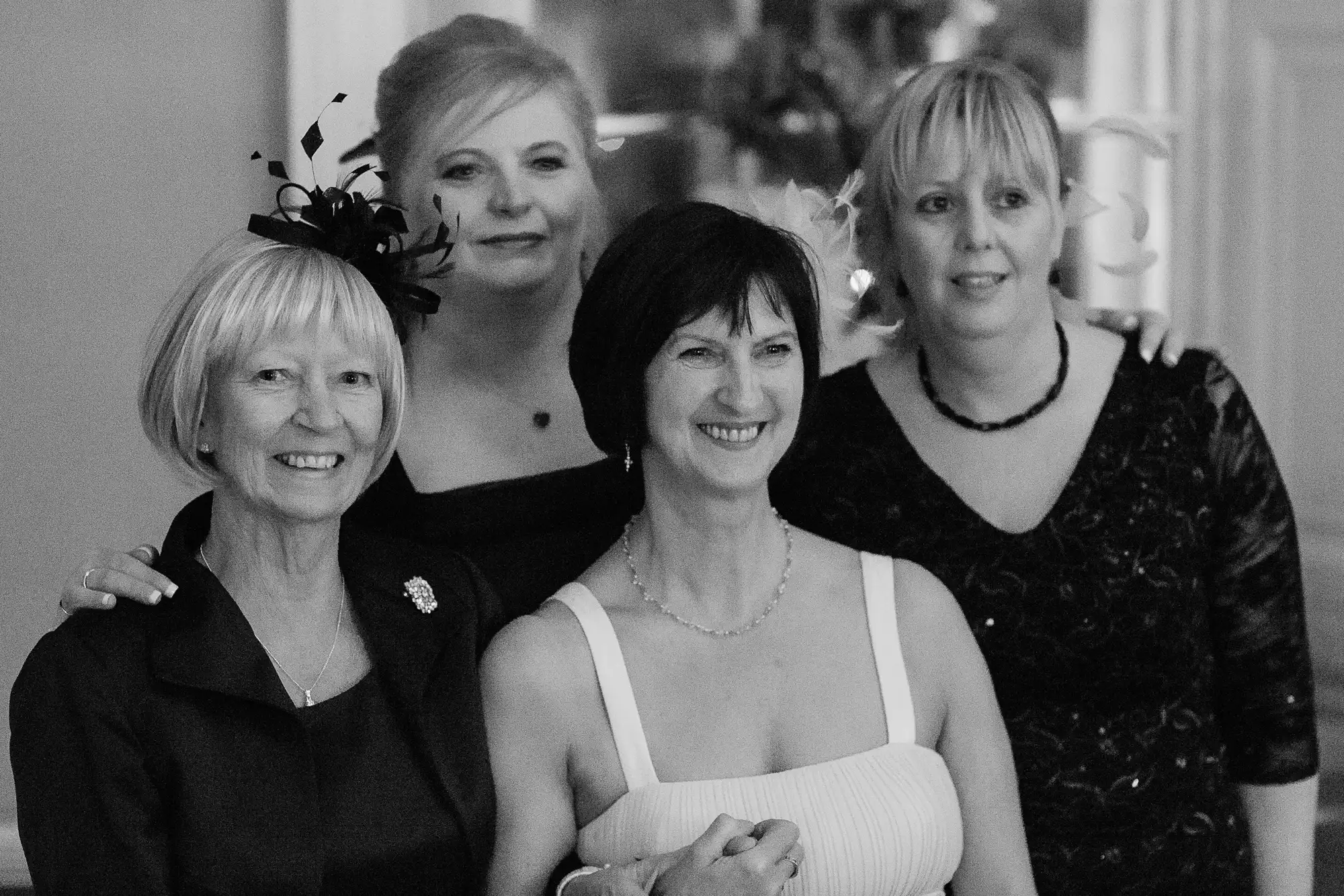 Four women smiling in a group, dressed in formal attire for a black and white photo.