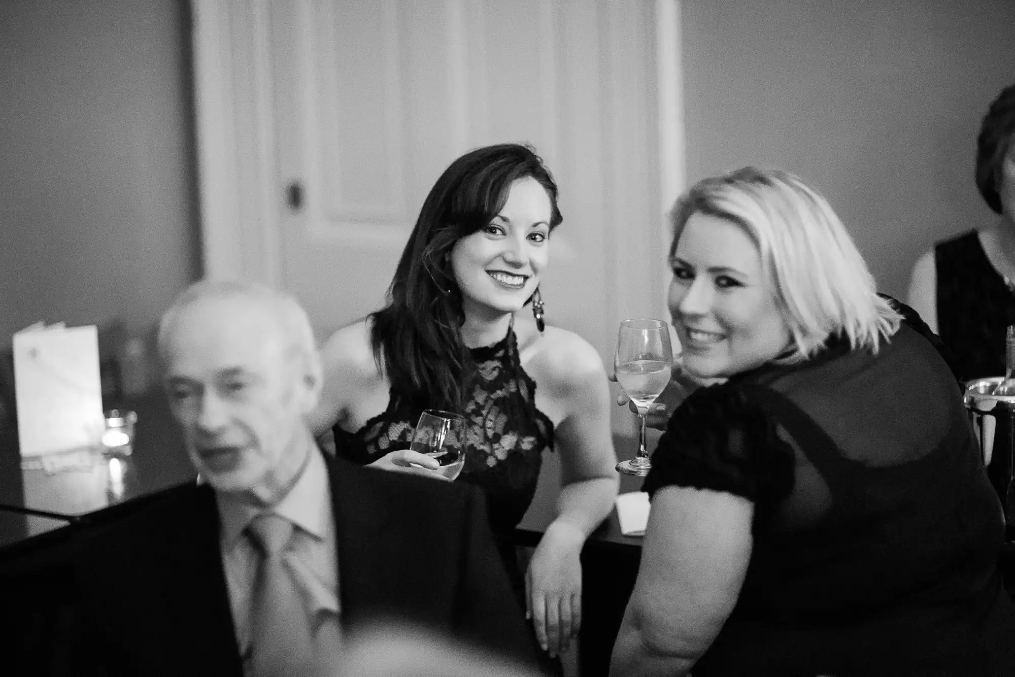 Two women smiling and sitting at a table during a formal event, with a blurred man in the foreground.