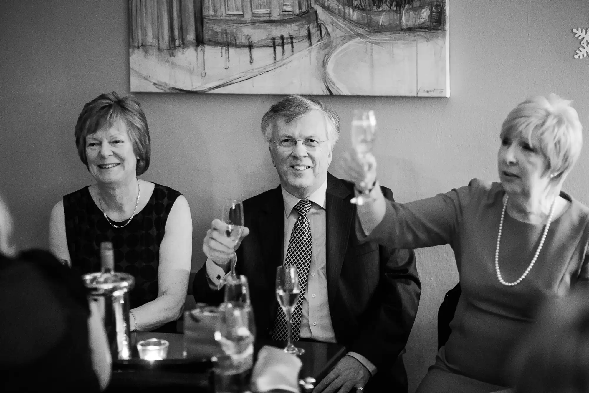 Three older adults enjoying a toast in a restaurant, with a painting in the background; two women and one man, all smiling.