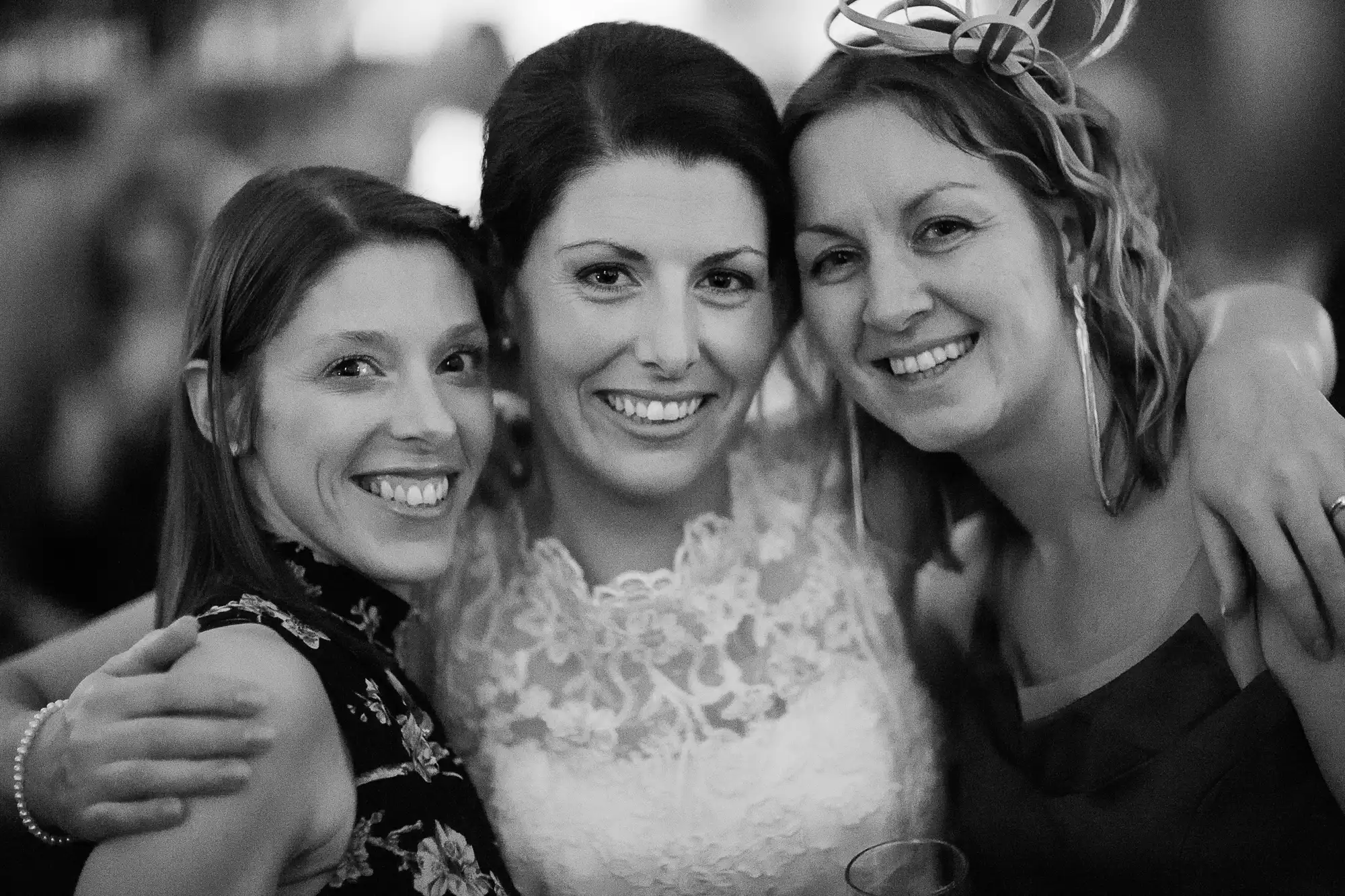 Three smiling women embracing at a festive event, one wearing a decorative headpiece, in a black and white photograph.