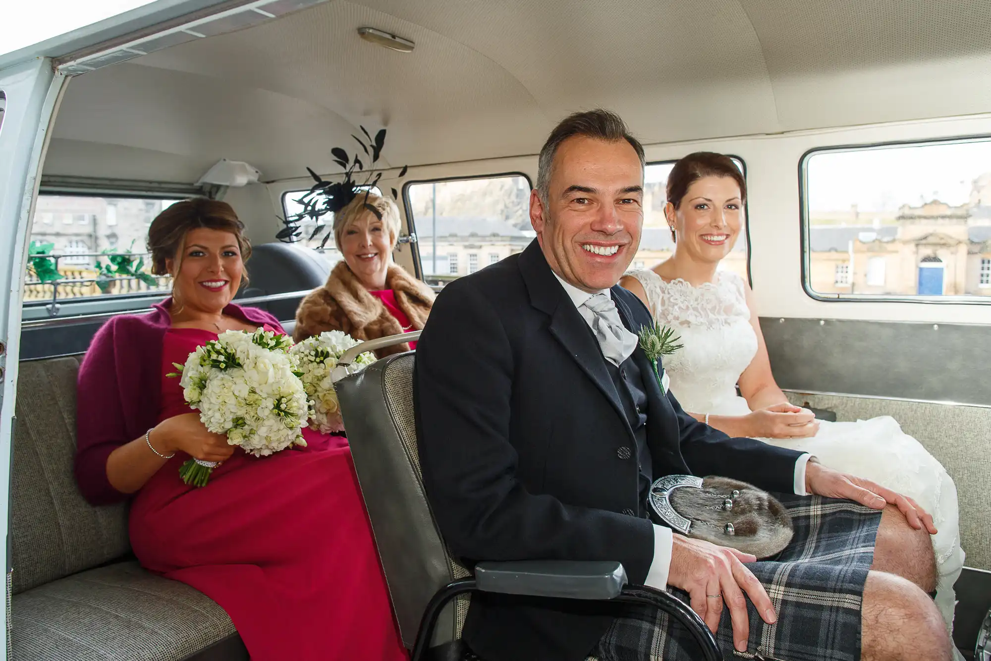 Bride, groom in kilt, and two female guests smiling inside a classic car, with floral bouquets and formal attire.