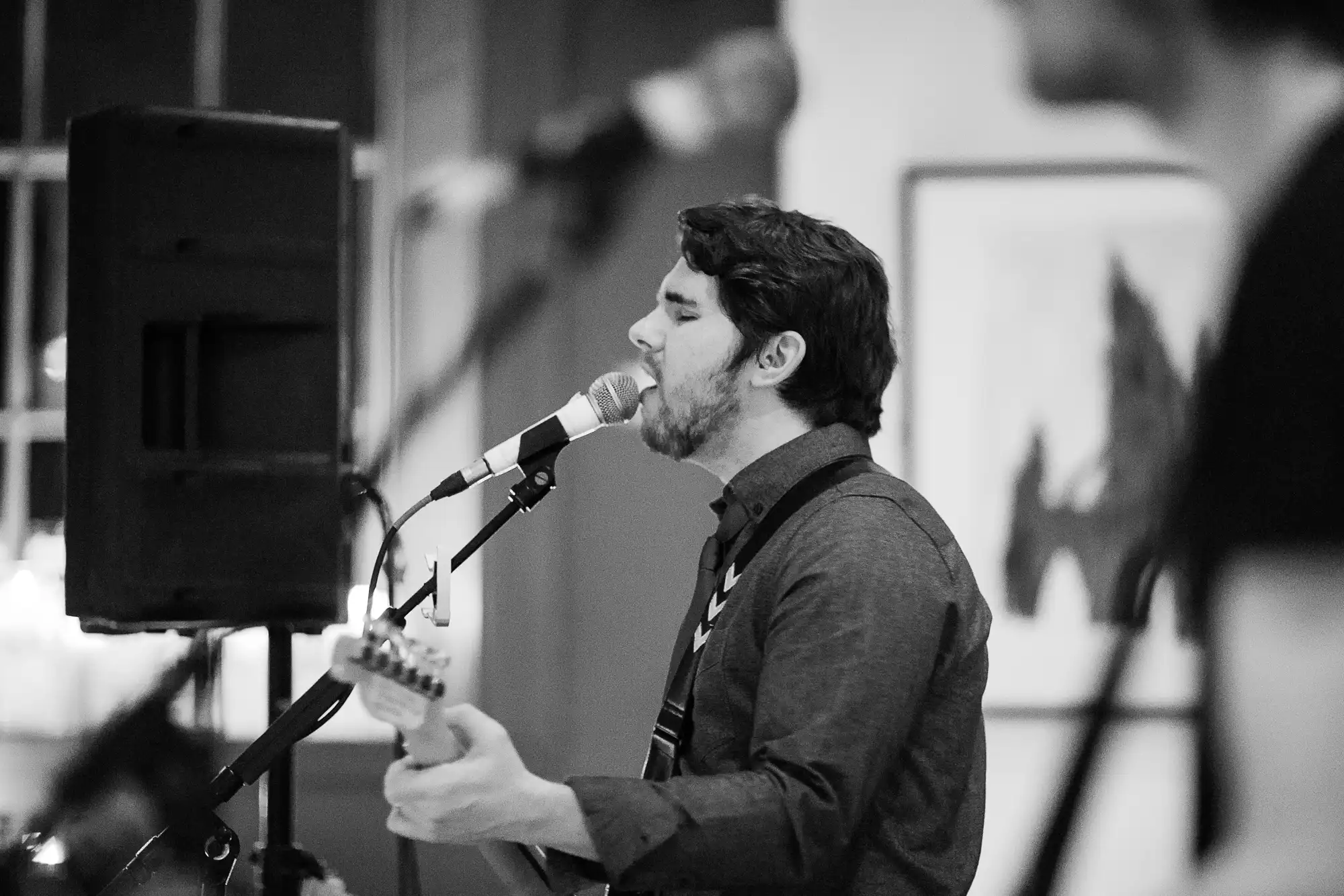 A man sings into a microphone while playing an electric guitar at an indoor event, in a black and white photo.