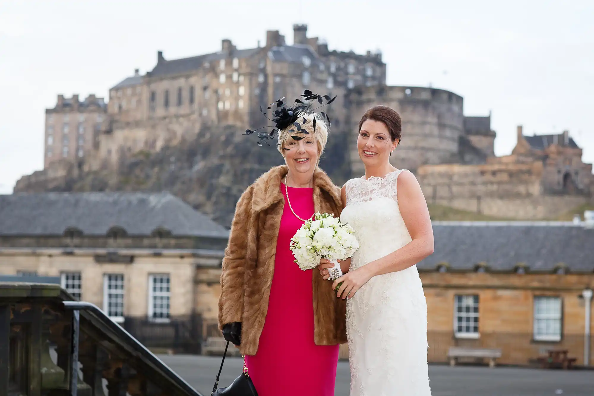 Two women, one in a wedding dress and the other in a red dress and black hat, standing in front of edinburgh castle.