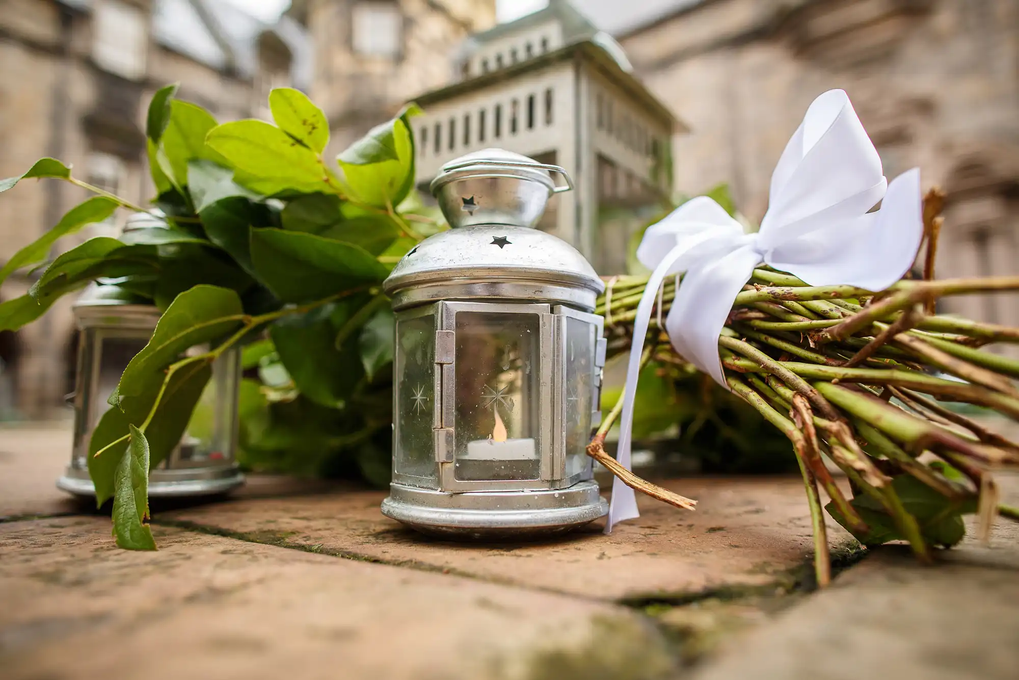 A metal lantern with a lit candle inside, surrounded by green leaves and a white bow, placed on a cobblestone surface.
