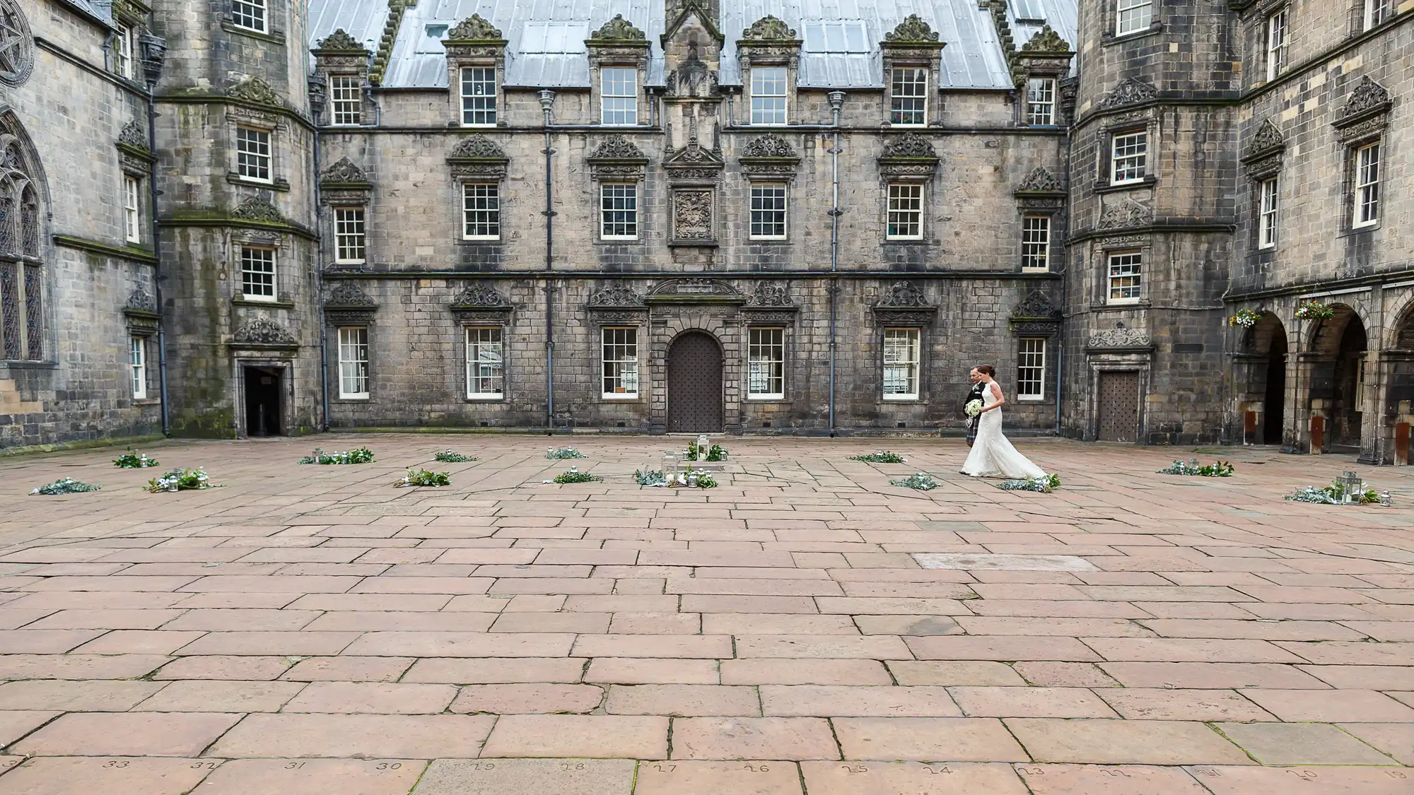 A bride walking alone across a cobblestone courtyard in front of an ornate, historical stone building with multiple windows and doors.