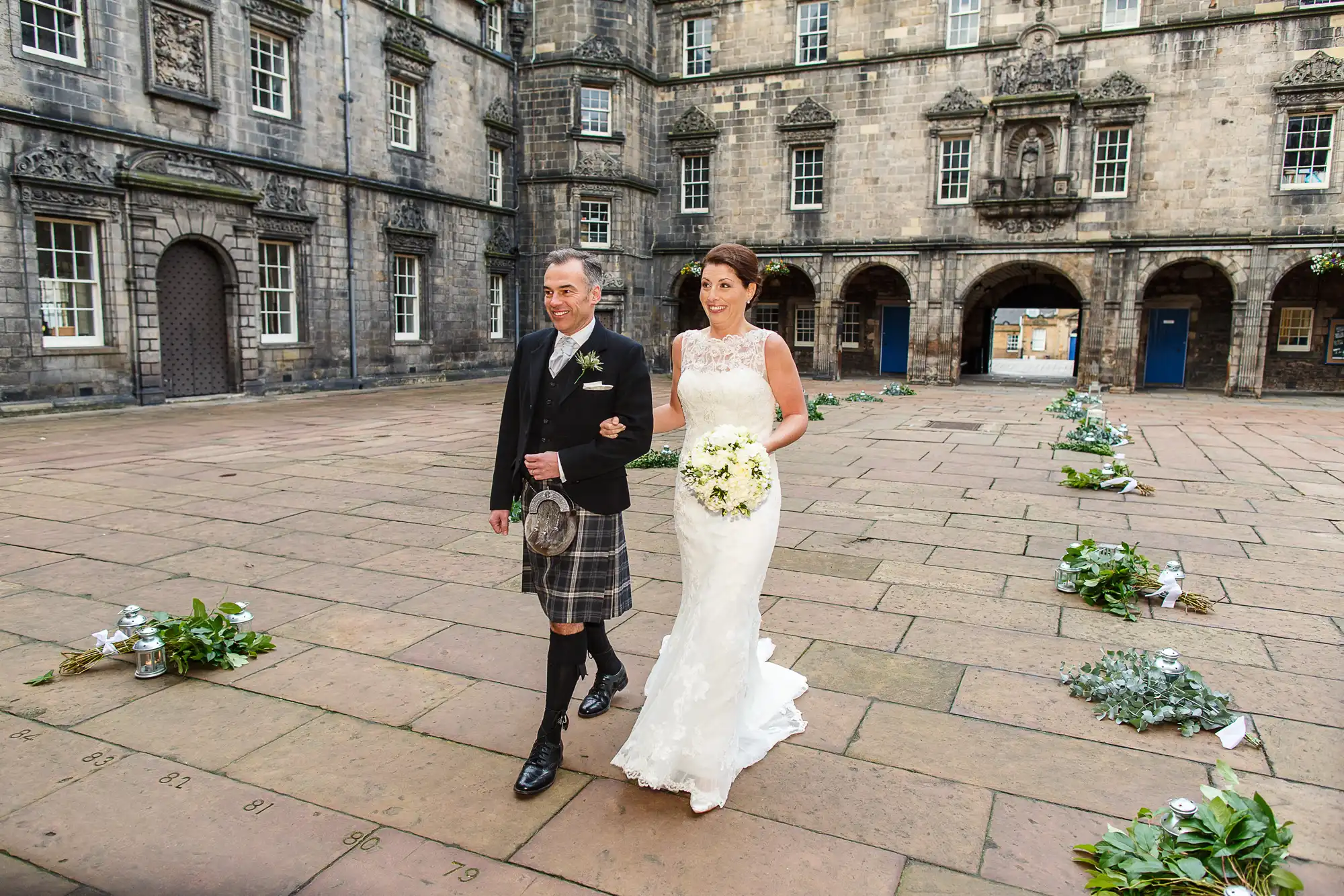 A bride in a white dress and a groom in a kilt and black jacket walk hand-in-hand across a courtyard with historic stone buildings in the background.