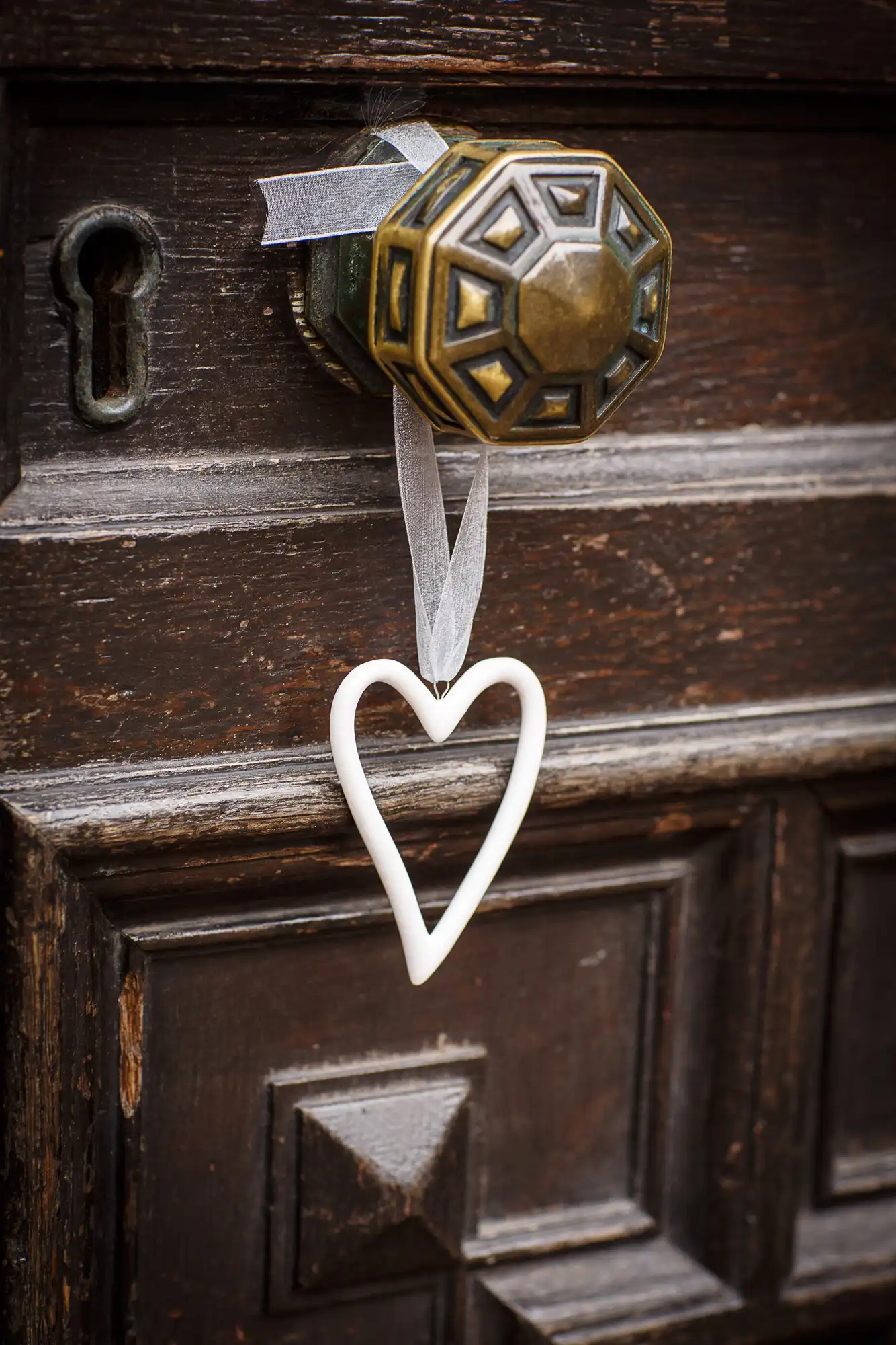 A white heart-shaped decoration hangs on a ribbon from an ornate brass door knob on an aged wooden door.