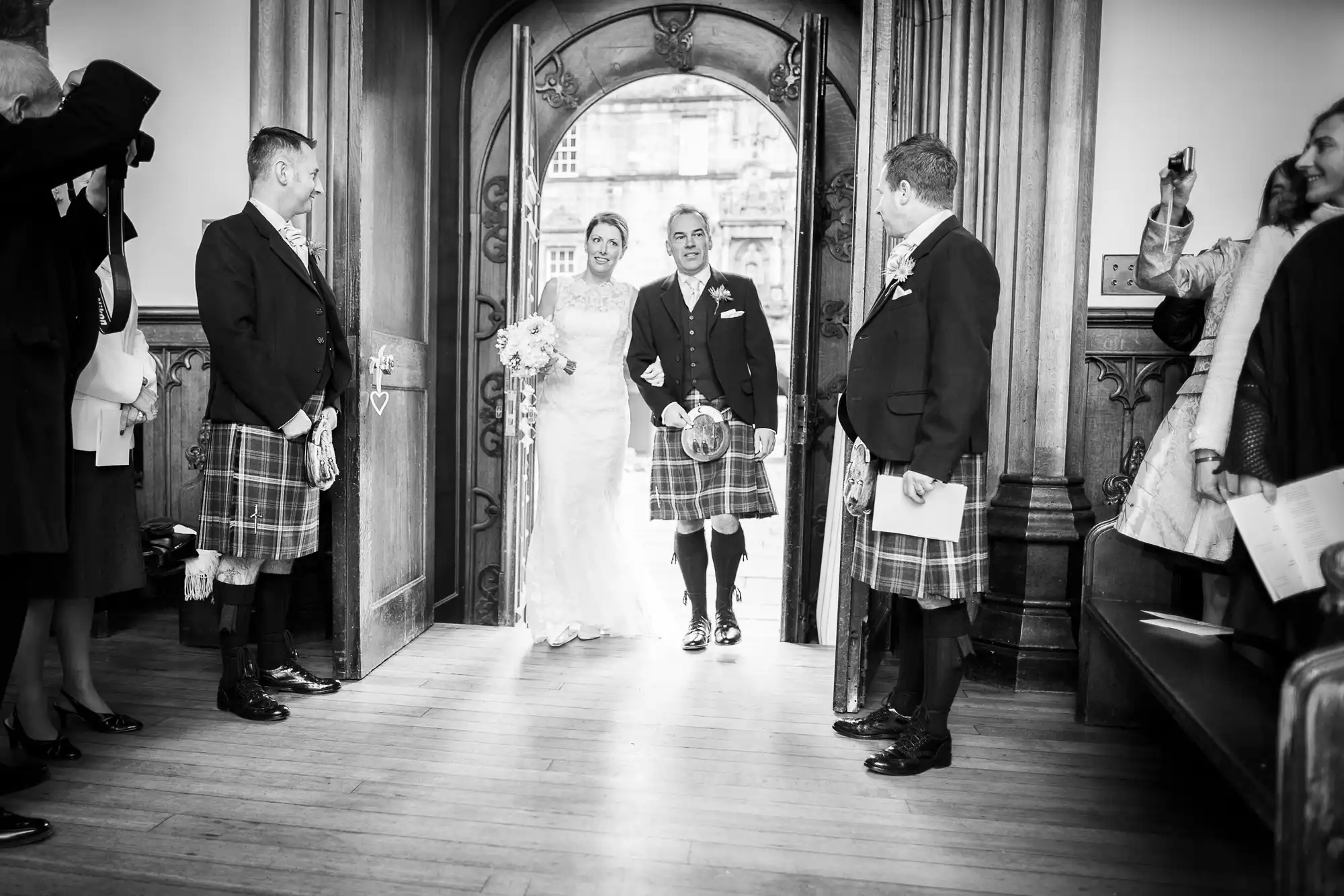 A bride and groom walk through a doorway smiling, surrounded by guests in kilts, at a traditional scottish wedding.