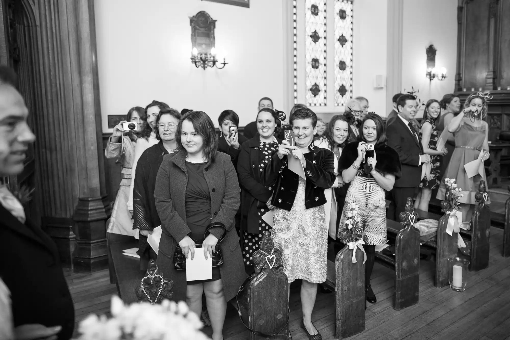 Guests smiling and clapping at a wedding ceremony inside a church hall, some holding cameras, captured in black and white.