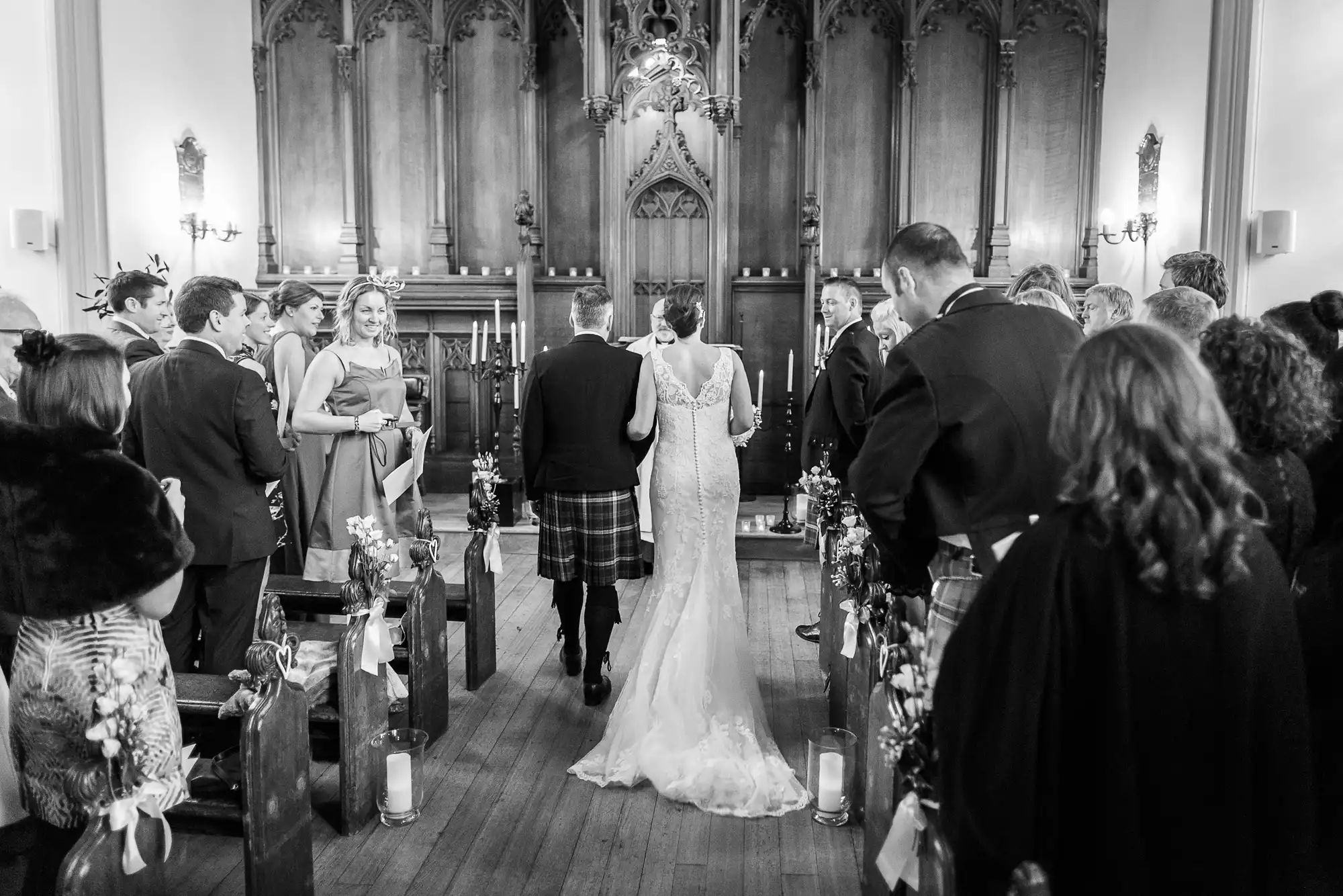 A bride and groom walk down the aisle of a church filled with guests during their wedding ceremony, captured in black and white.