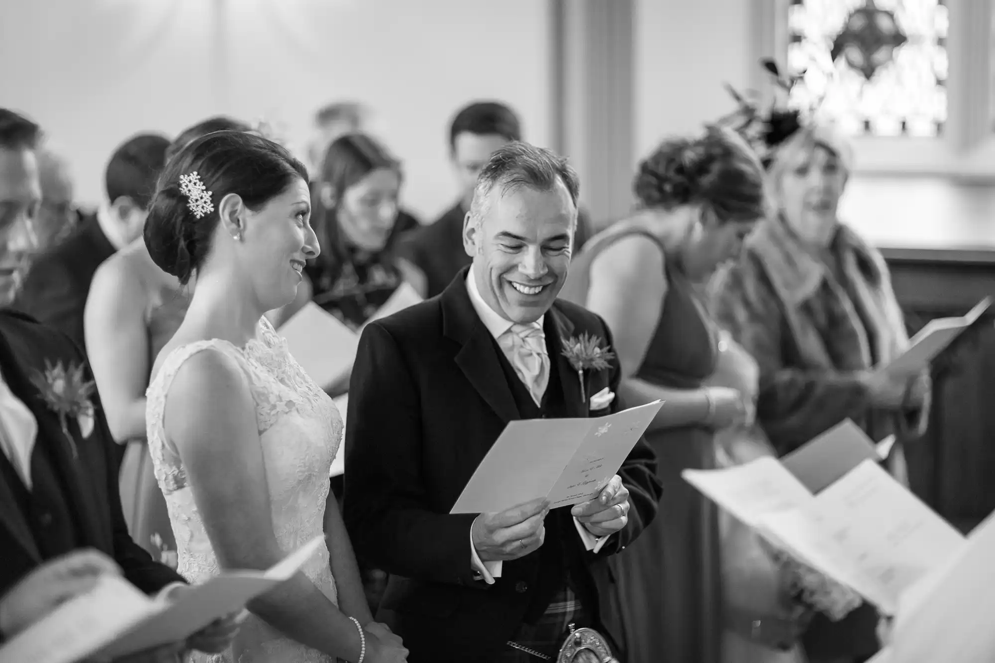 A bride and groom smiling and looking at a document during their wedding ceremony, surrounded by guests in a black and white photo.