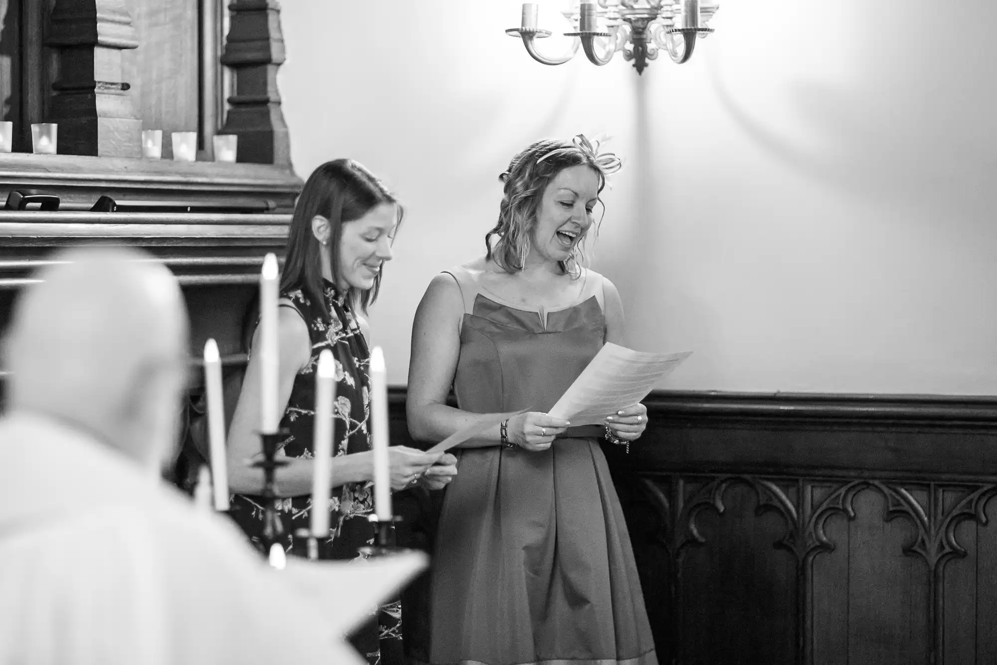 Two women standing and laughing together in a church, one holding a sheet of paper, in a black and white photo.