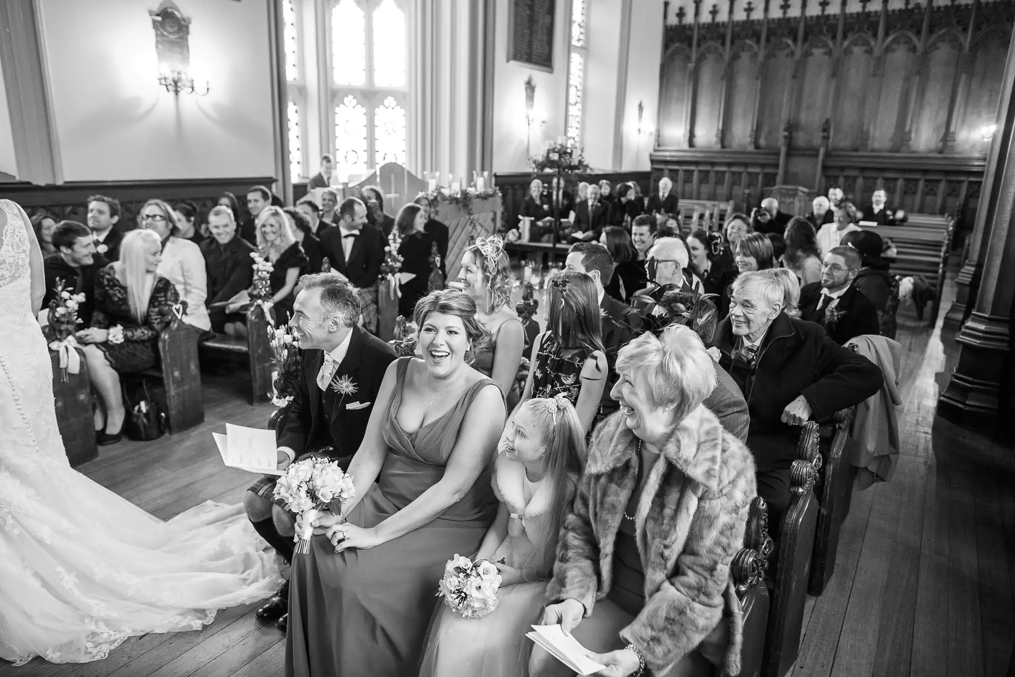 Black and white photo of guests seated at a wedding ceremony inside a church, smiling as they watch the bride standing to the left.