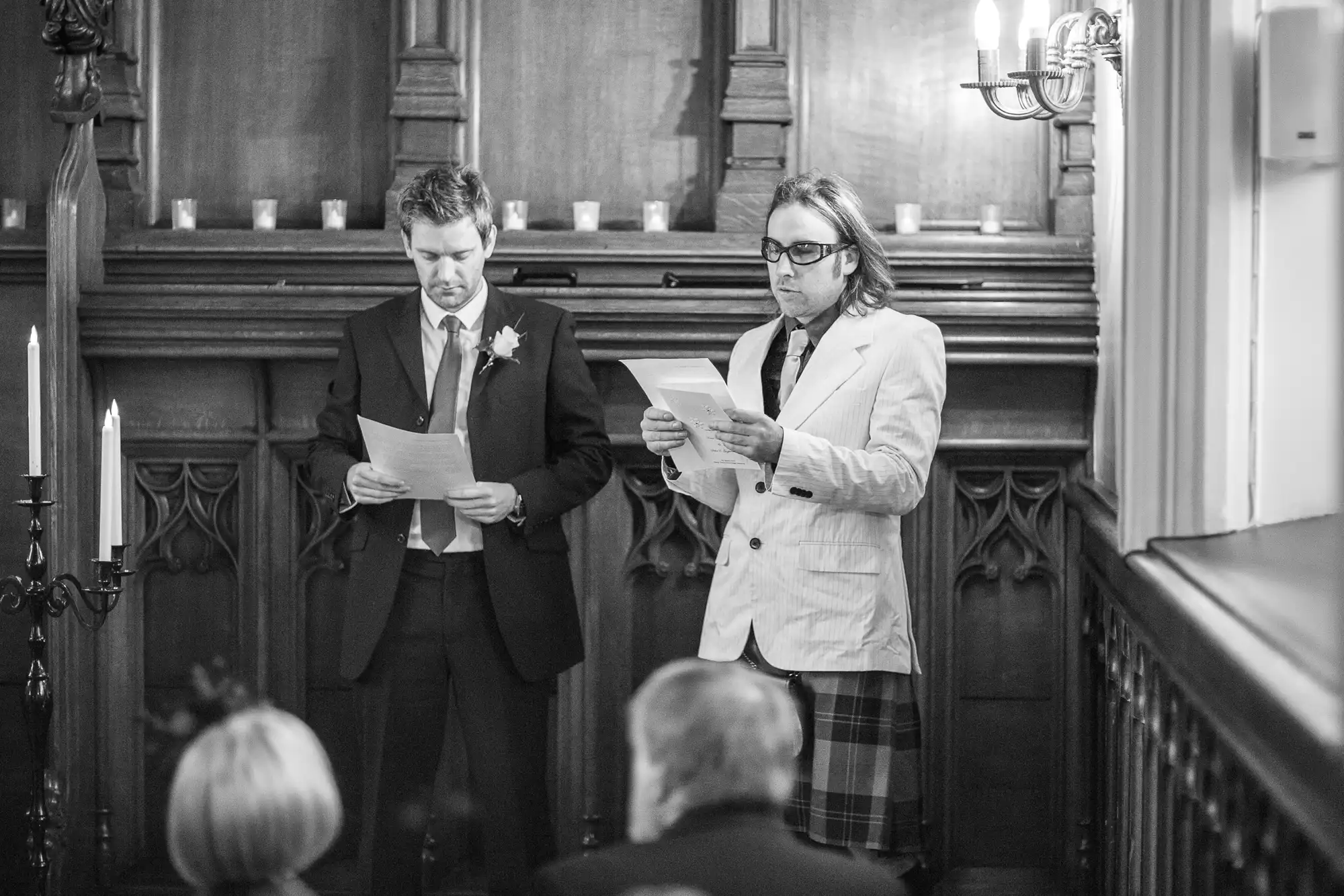 Two men reading from papers at a formal event inside a church, with one man in a traditional kilt and the other in a suit.