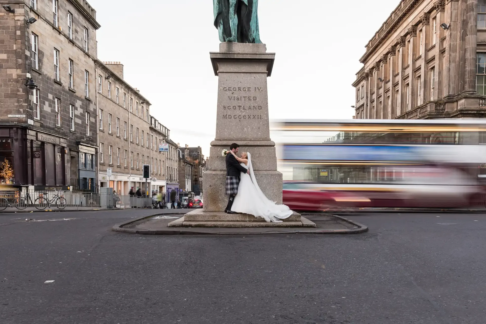 A newlywed couple poses for a photo at the base of a George IV statue, with a blurred bus passing by in the background.