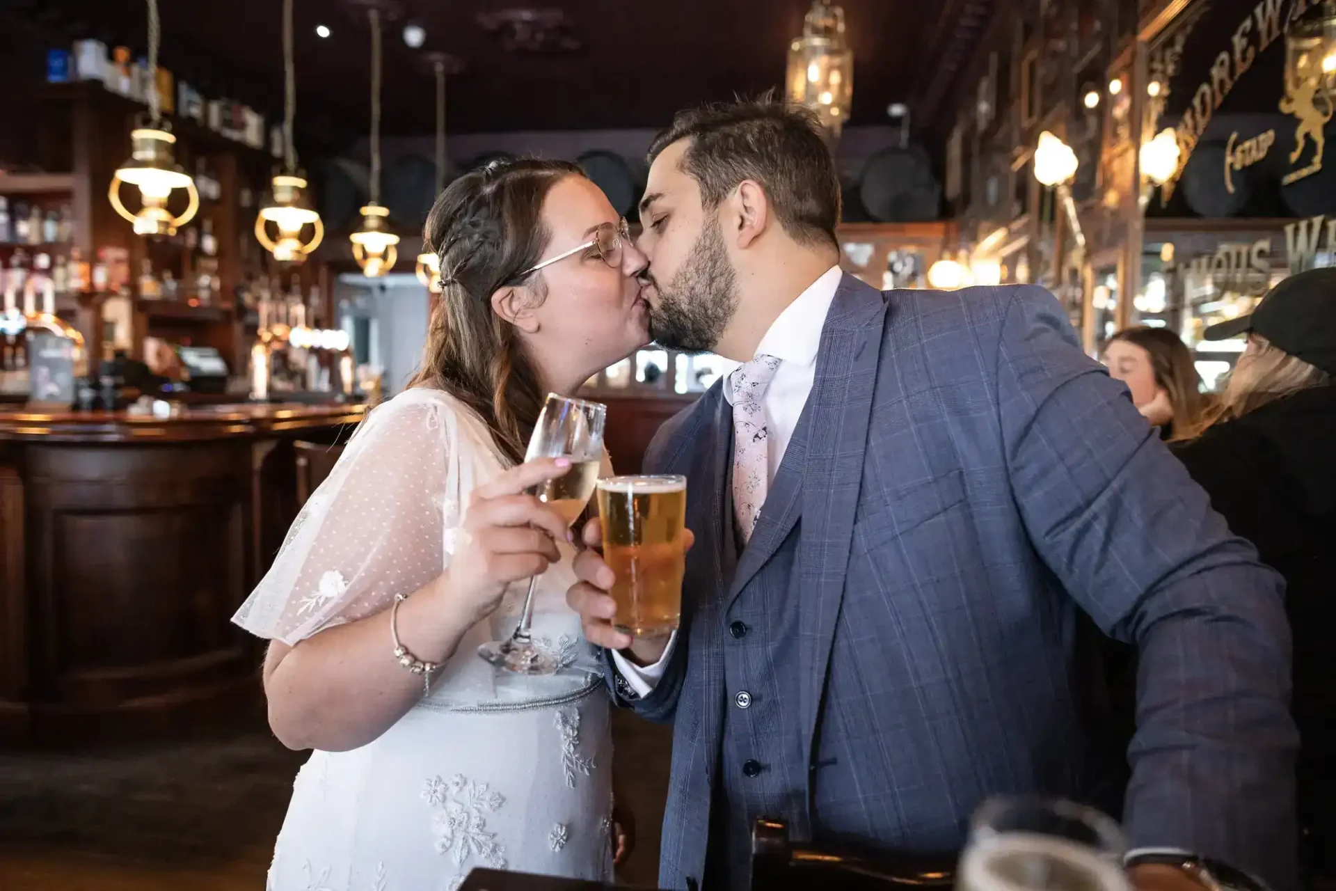 A couple dressed in formal attire kiss while holding drinks, a glass of beer and a glass of wine, inside a pub with dark wood and warm lighting.