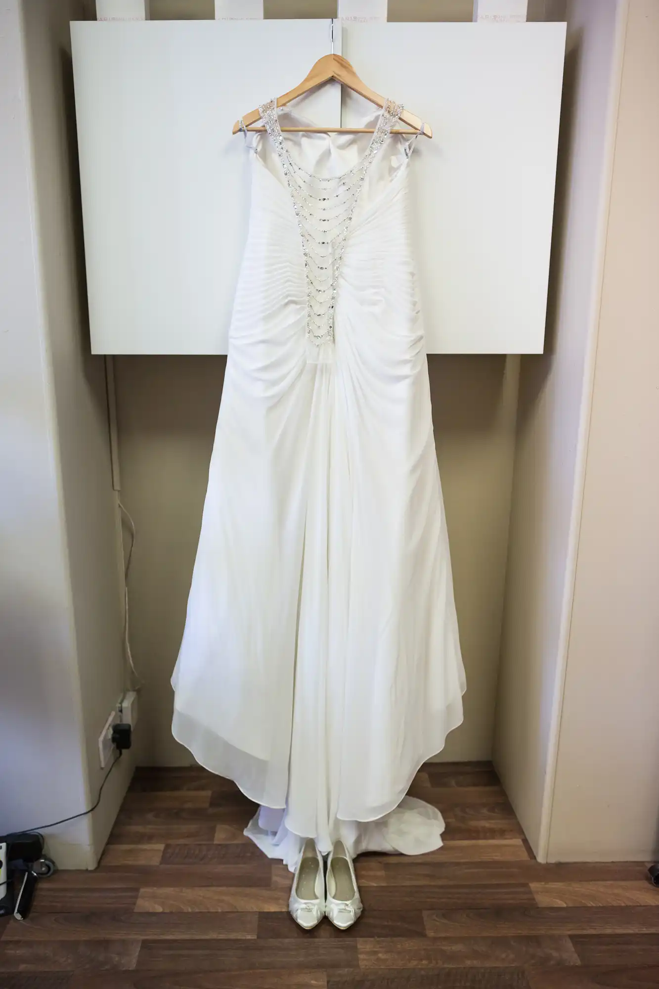 A white wedding dress with detailed beadwork on the bodice hangs on a wooden hanger against a blank canvas, with white shoes placed below it on a wooden floor.