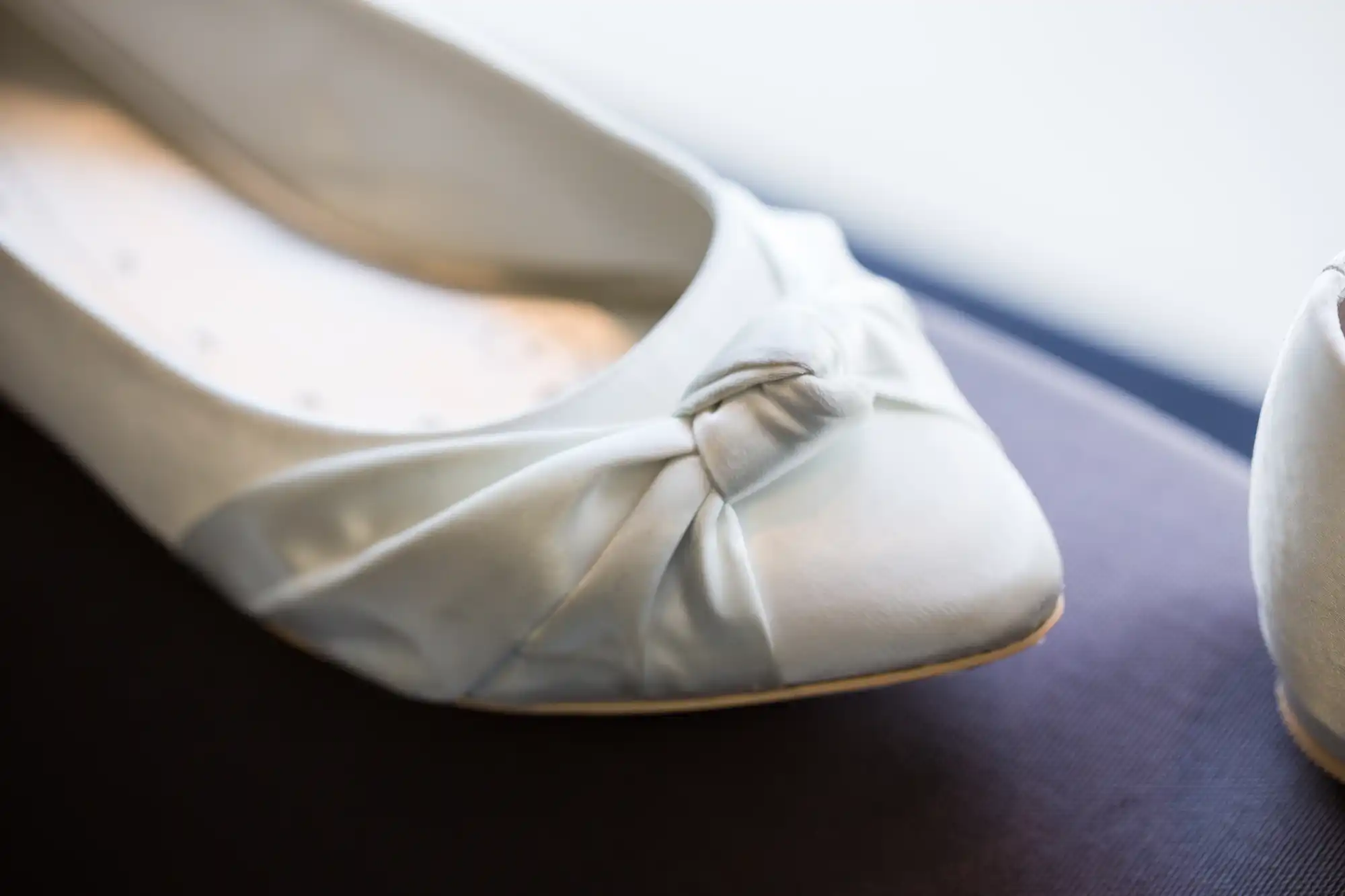 Close-up of a worn white ballet flat with a twisted bow on the toe, resting on a dark surface.