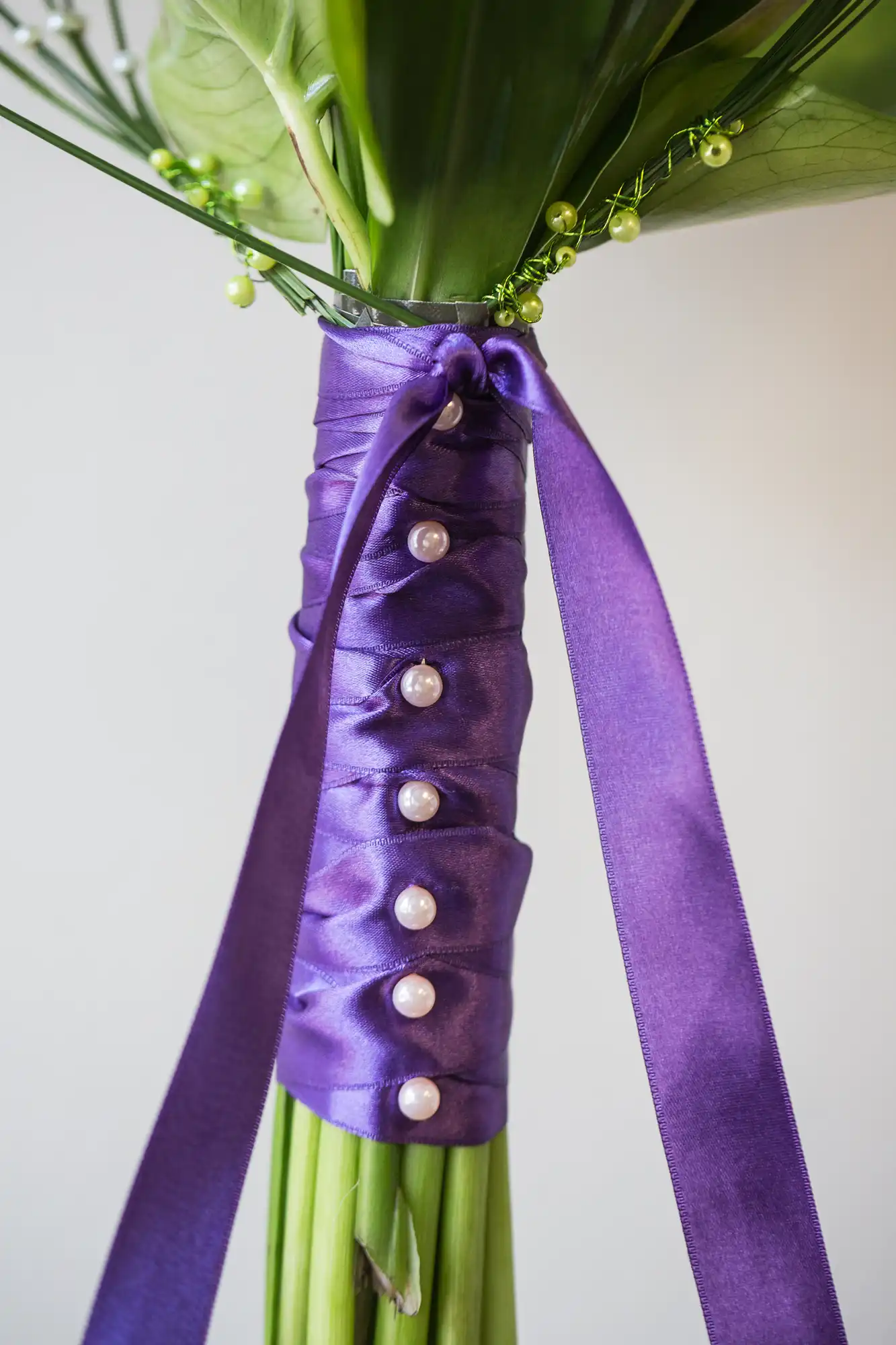 A close-up of a bouquet handle wrapped in purple satin ribbon adorned with pearls, with green stems visible.