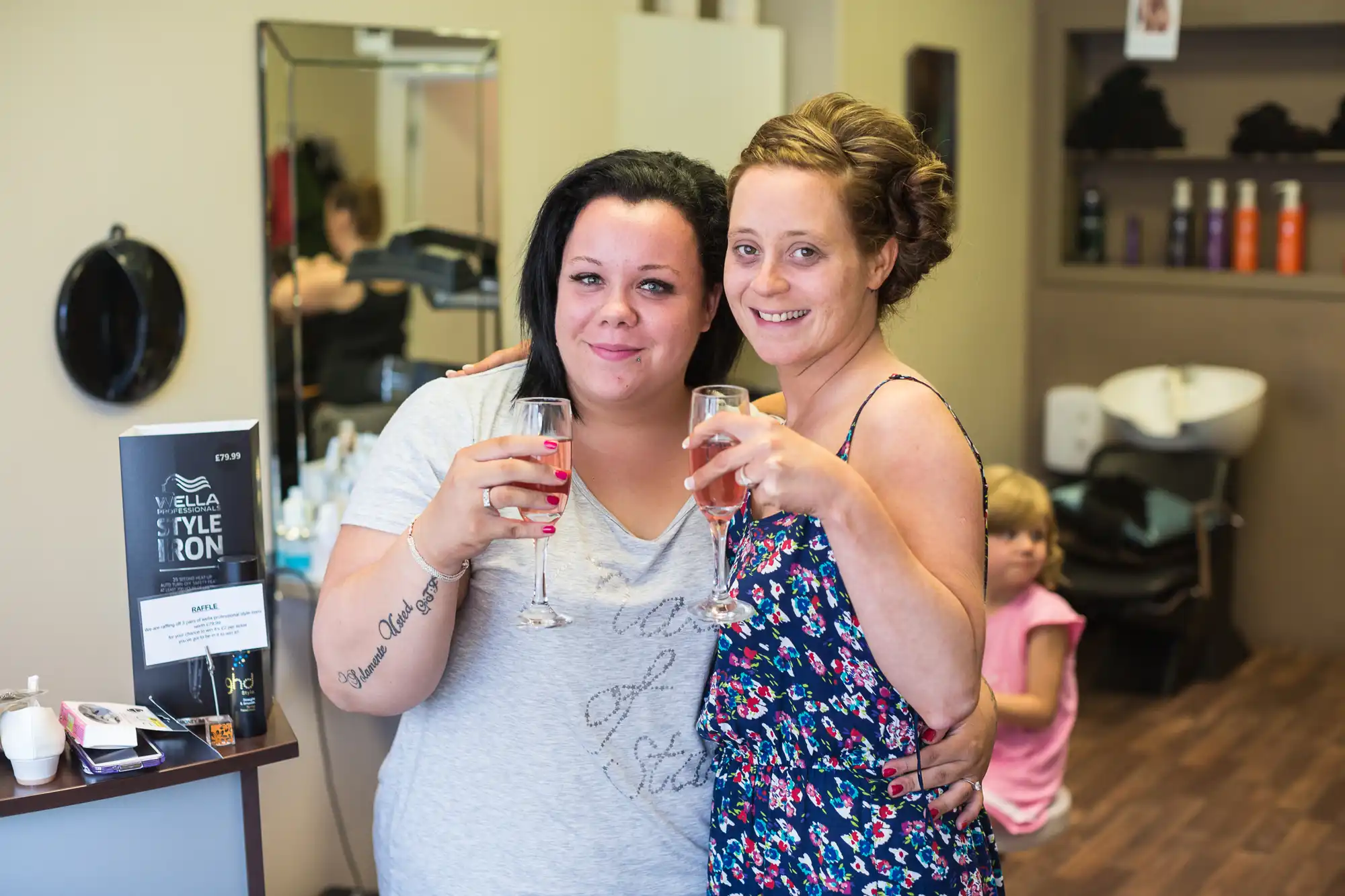 Two women smiling and toasting with glasses of wine in a hair salon, with a child visible in the background.