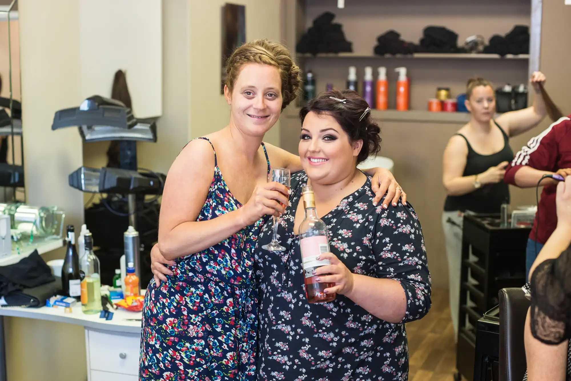 Two women smiling and holding champagne glasses in a busy salon, with other people and hairstyling equipment in the background.