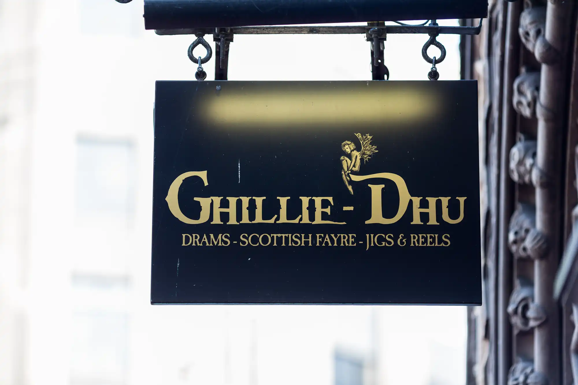 Black and gold sign reading "ghillie-dhu - drams - scottish fayre - jigs & reels" hanging on metal chains, with a blurred building background.
