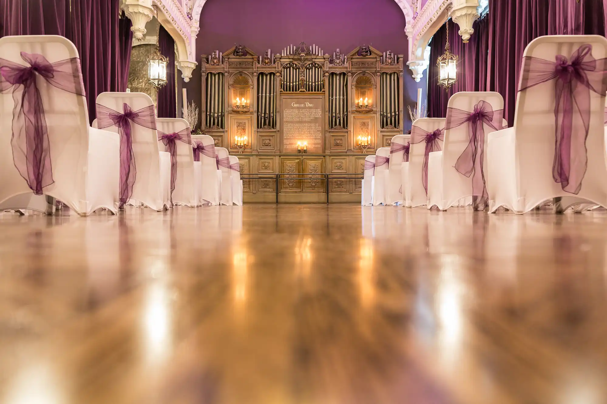 Elegant wedding venue interior with white chairs adorned with purple ribbons and a large, ornate organ at the front illuminated by soft lighting.