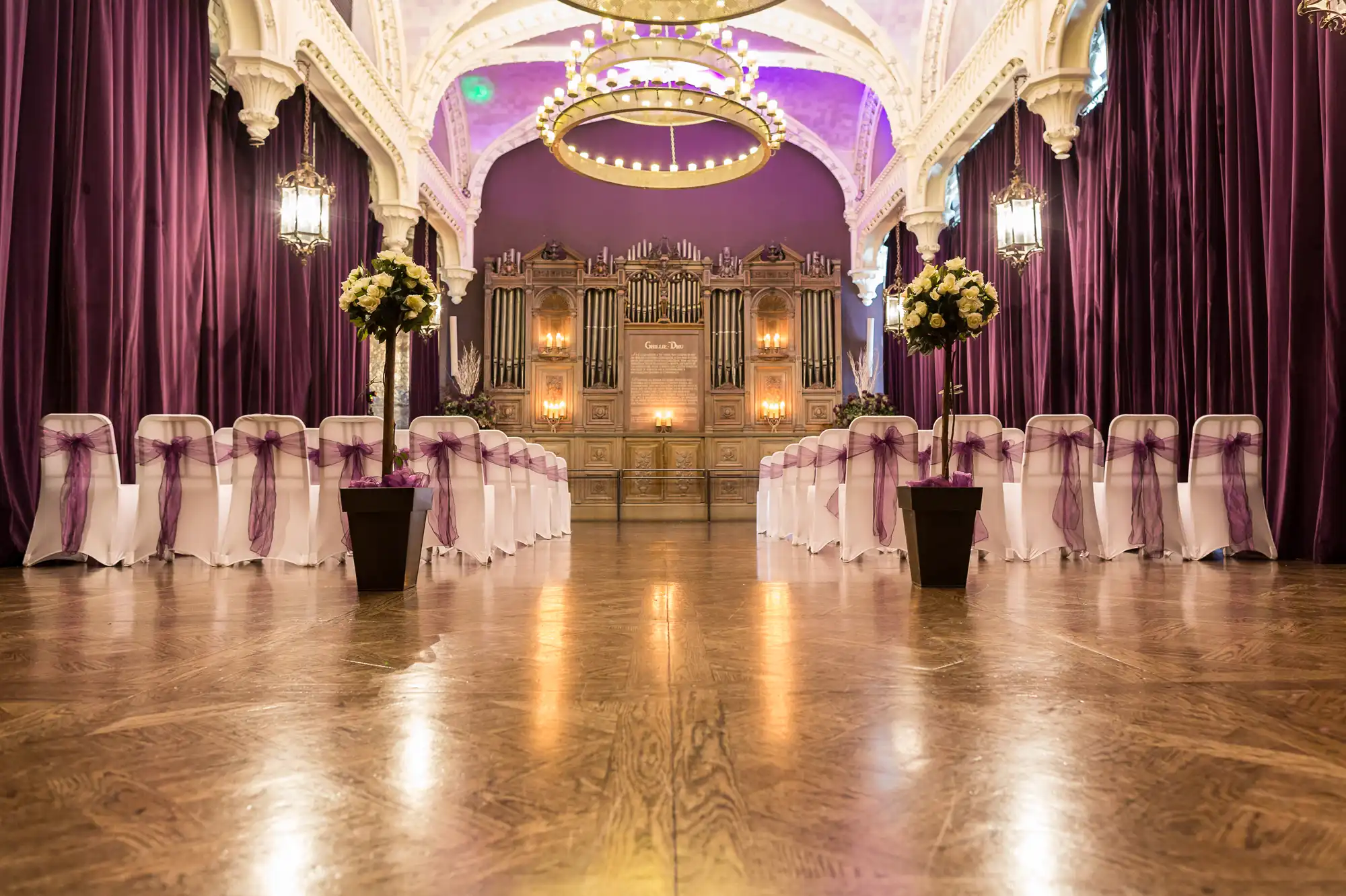 Elegant wedding venue interior with a purple color scheme, white chairs covered with purple ribbons, floral decorations, and a grand chandelier.