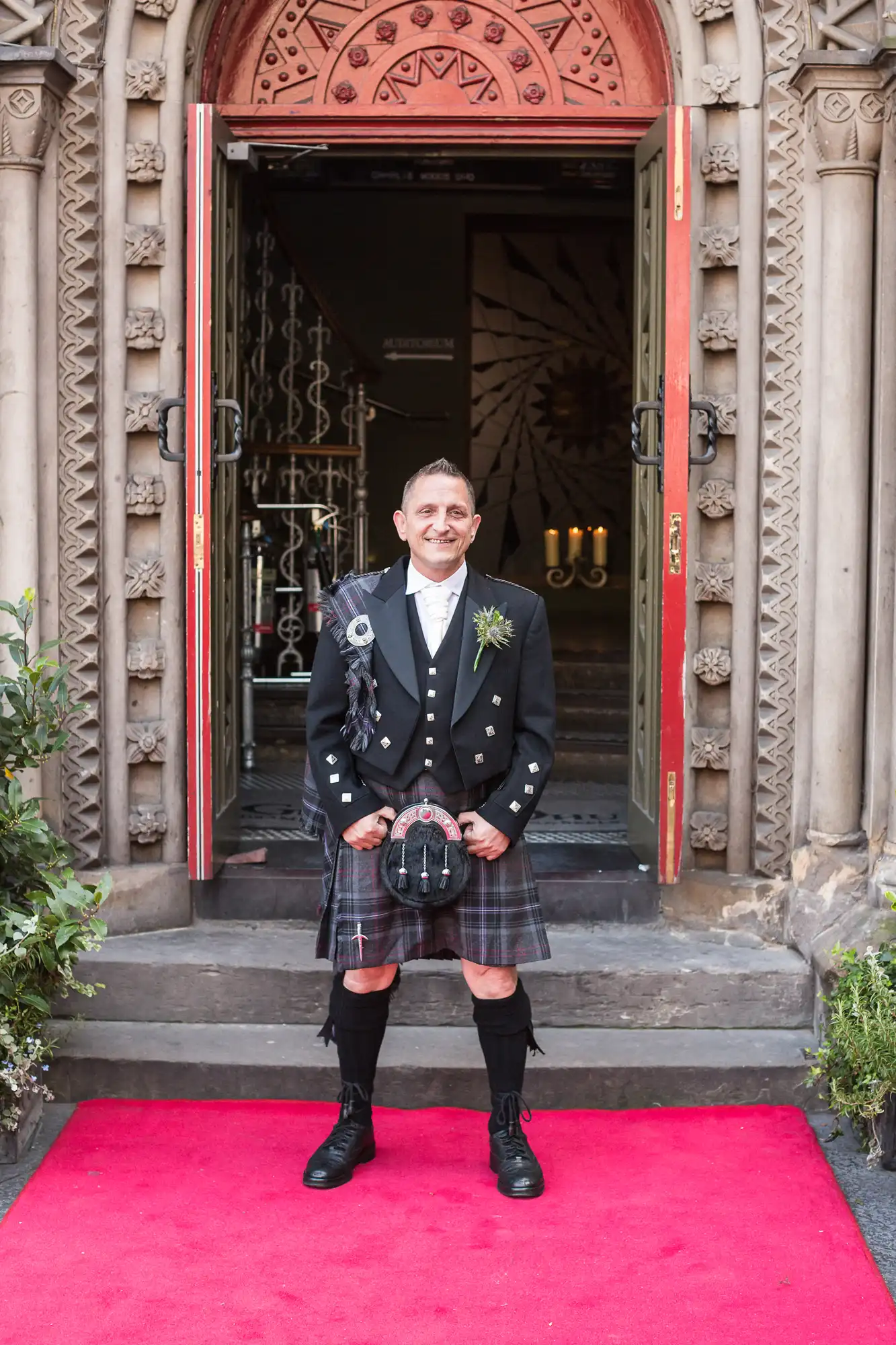 A smiling man in traditional scottish attire, including a kilt and sporran, stands on a red carpet at an ornate doorway.